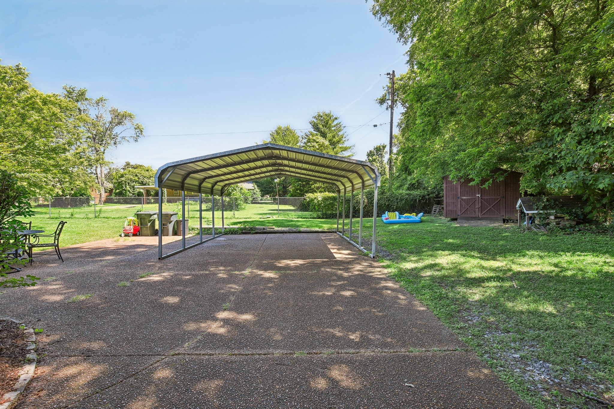 Backyard driveway with a large metal carport, wooden shed, and leafy lawn.