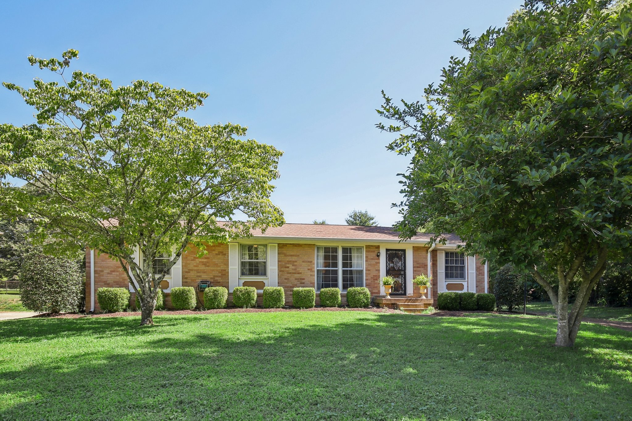 Single-story brick ranch house with white shutters, clipped shrubs, and a shady front lawn.
