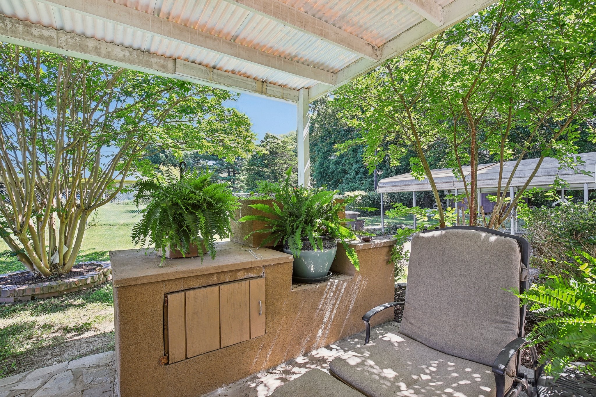 Covered patio with a built-in counter, potted ferns, and a cushioned chair beneath leafy trees.