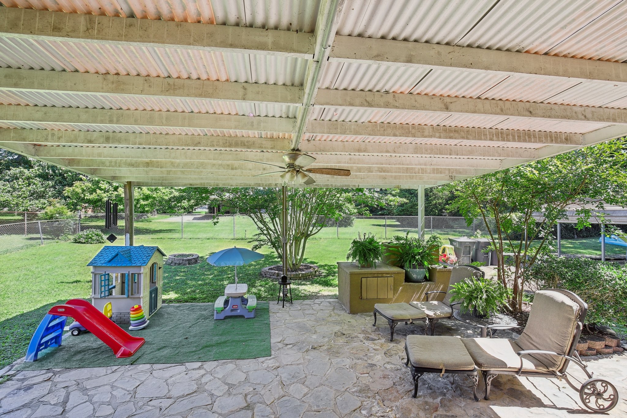 Covered stone patio with lounge chairs and play equipment beside a fenced, tree-lined lawn.
