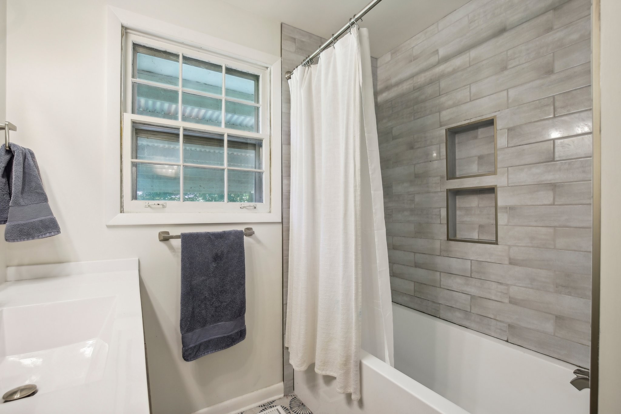 Bathroom with a gray tiled tub and shower, two inset niches, and a large window.