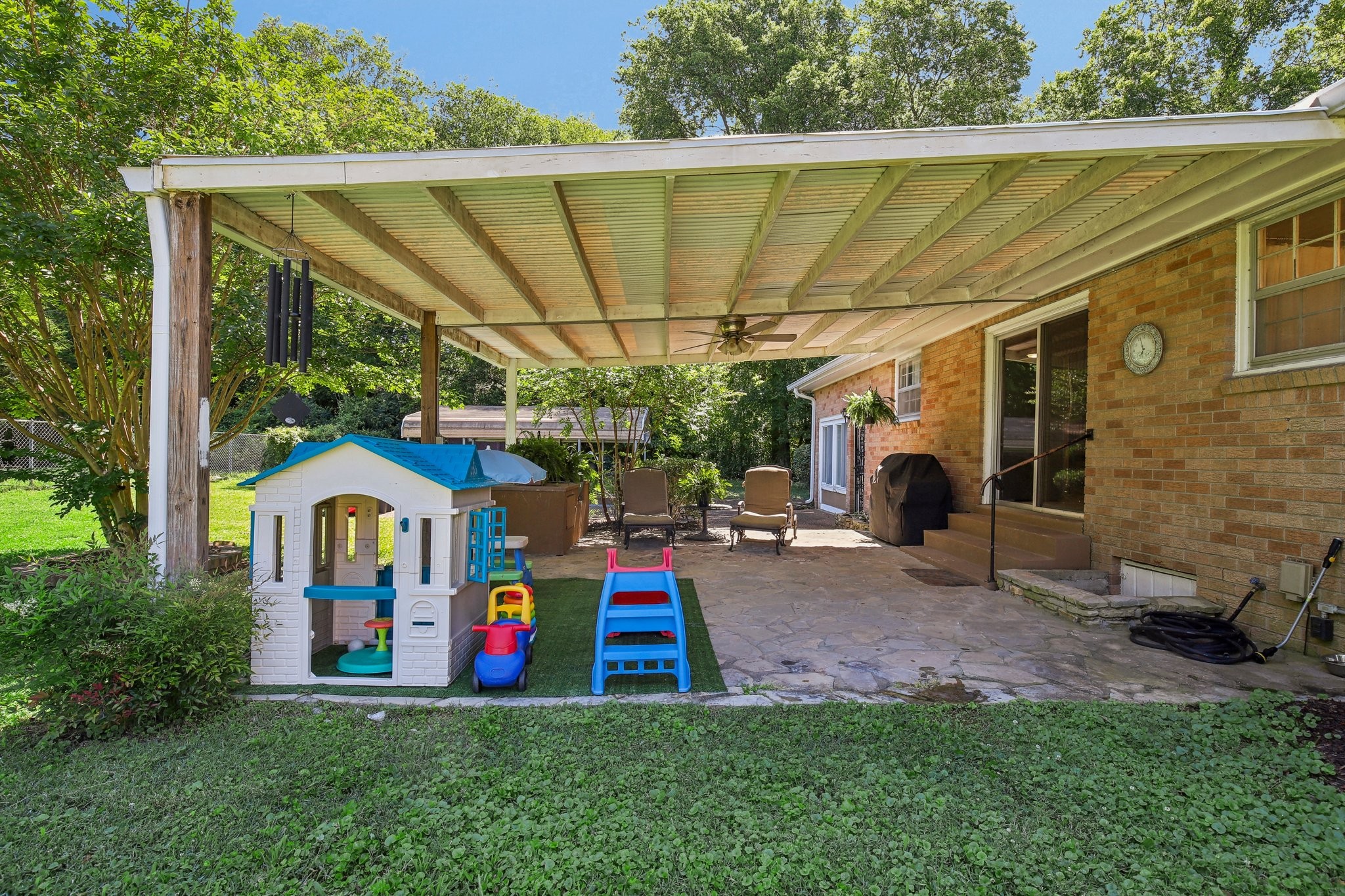 Covered backyard patio with a stone floor, outdoor chairs, and children’s play equipment.