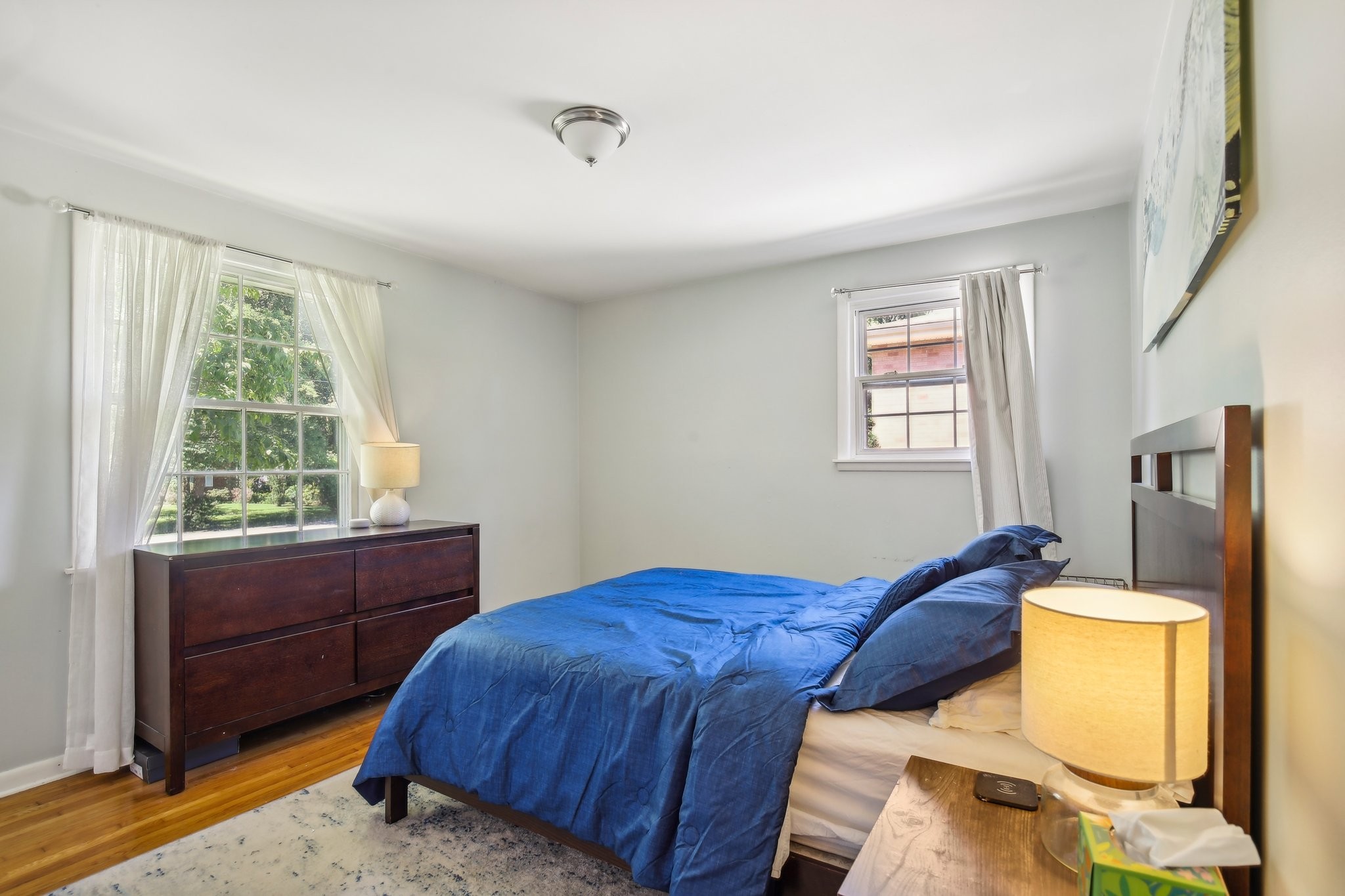 Bedroom with blue bedding, a dark wood dresser, hardwood floors, and two curtained windows.