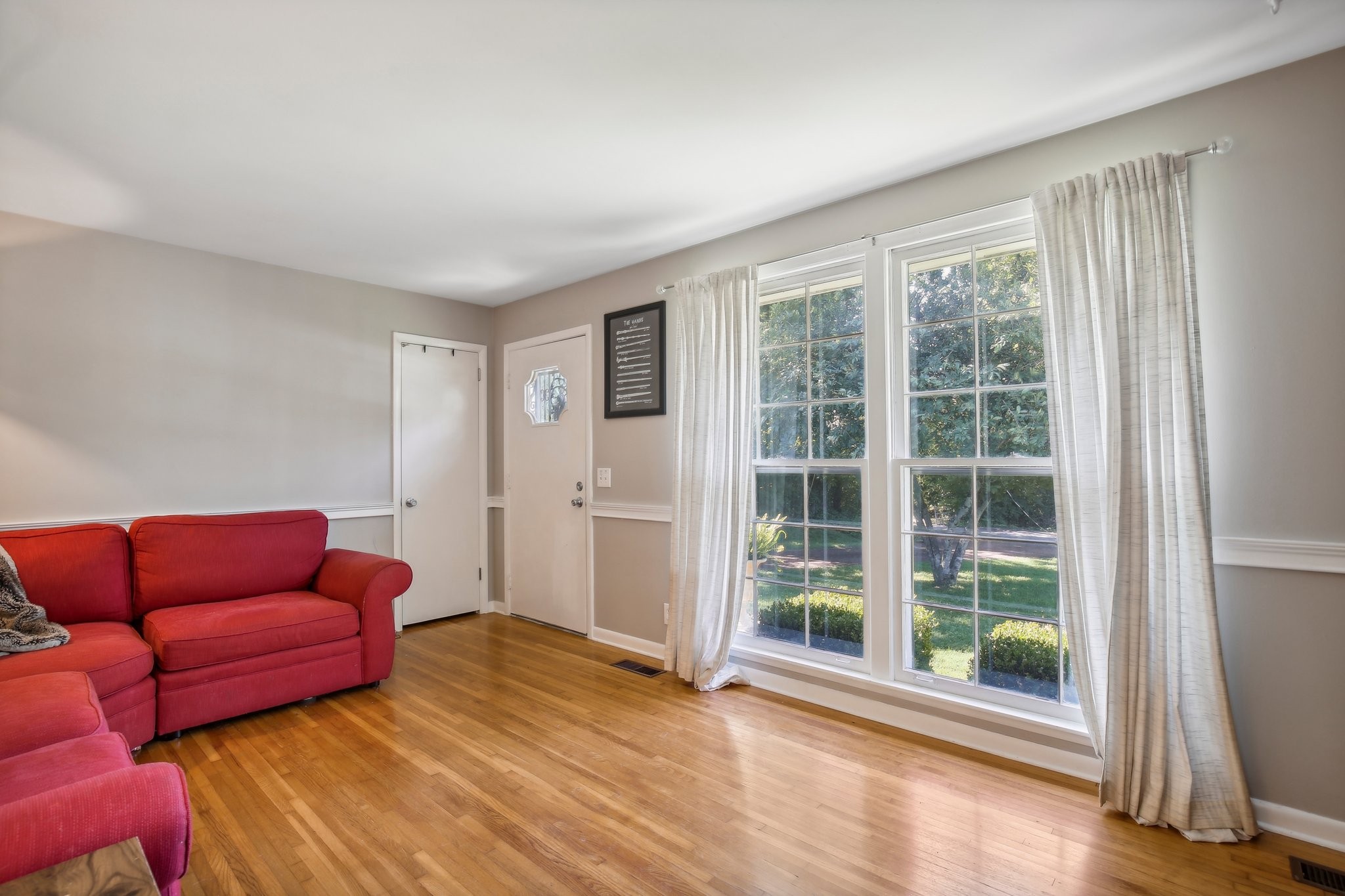 Living room with a red sectional sofa, hardwood floors, and tall windows overlooking a leafy yard.