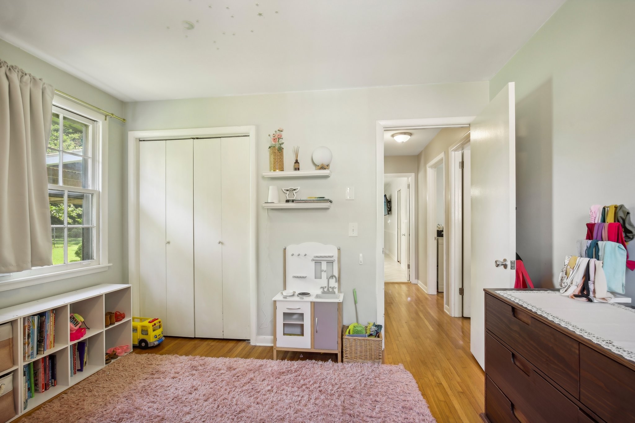 Child’s bedroom with a pink rug, toy kitchen, bookshelves, and a window overlooking the yard.