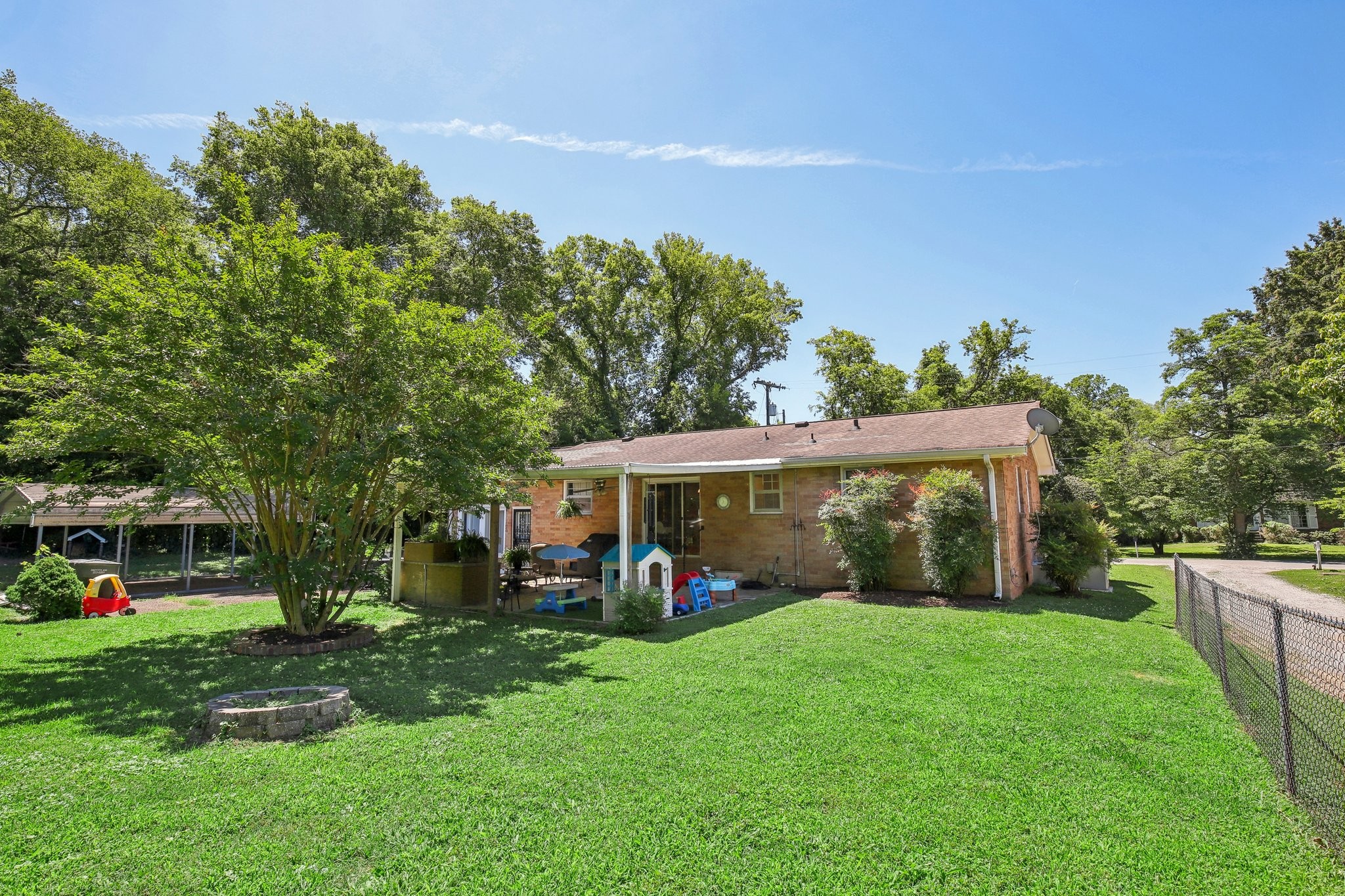 Brick home’s rear patio and broad lawn beneath mature trees, with children’s play equipment