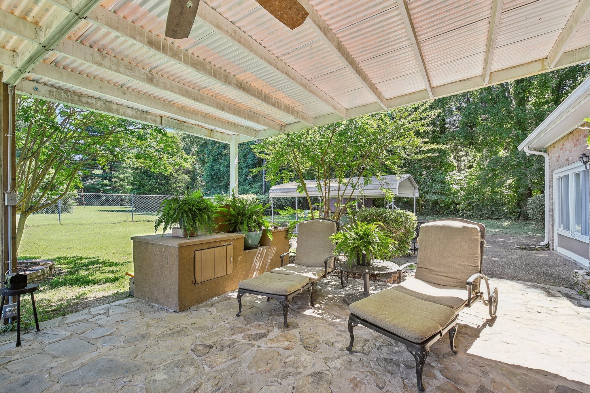 Covered stone patio with cushioned lounge chairs and ferns overlooking a green yard.