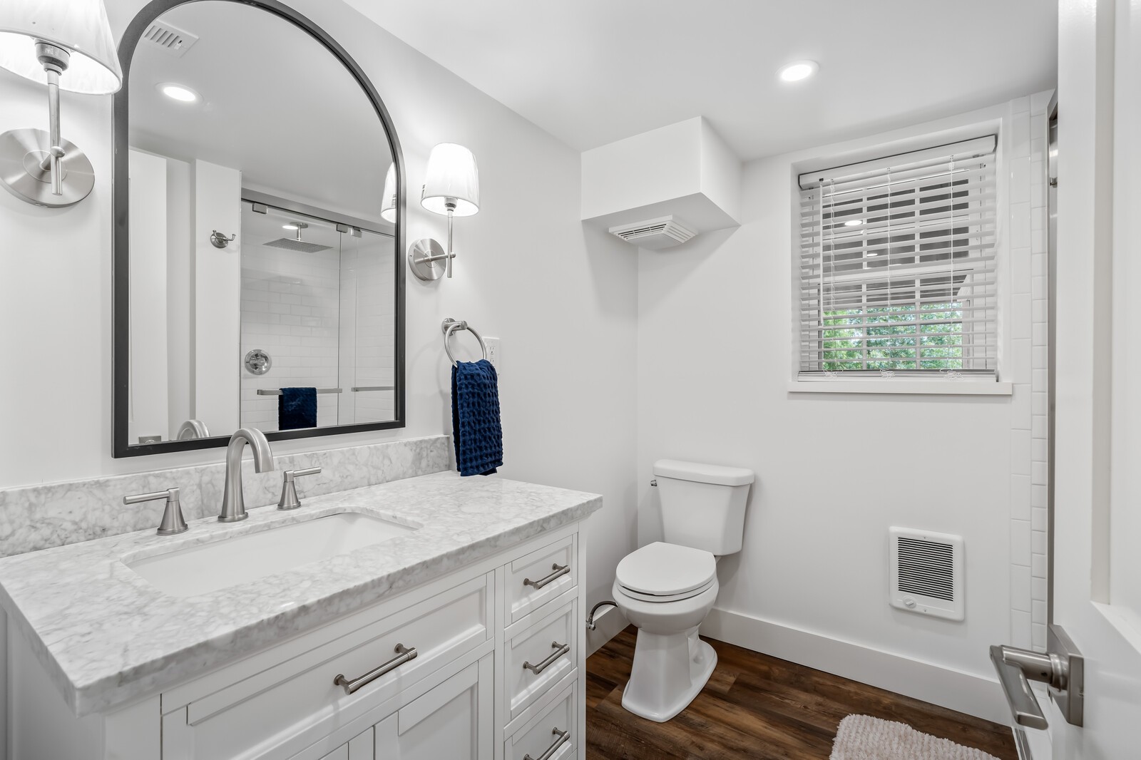 Bright bathroom with a marble-pattern vanity, framed mirror, glass shower, and window with white blinds.
