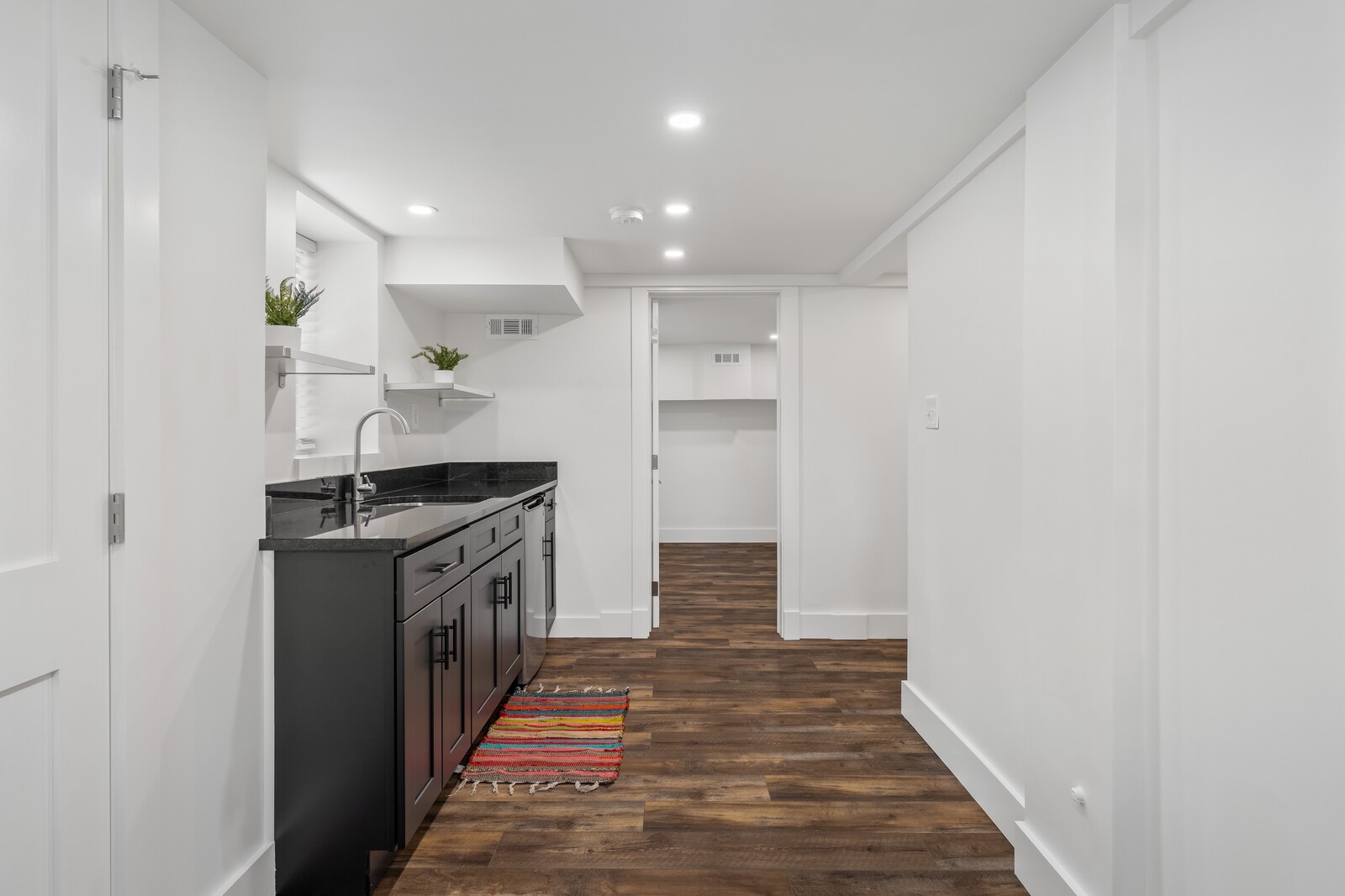Compact kitchenette with dark cabinets, black counters, floating shelves, and wood-look flooring.
