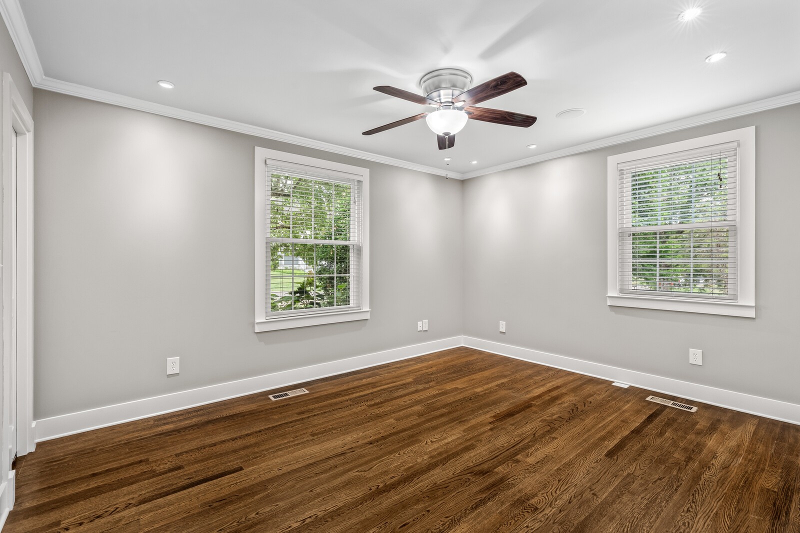 Empty room with hardwood floors, two windows, and a dark wood ceiling fan.