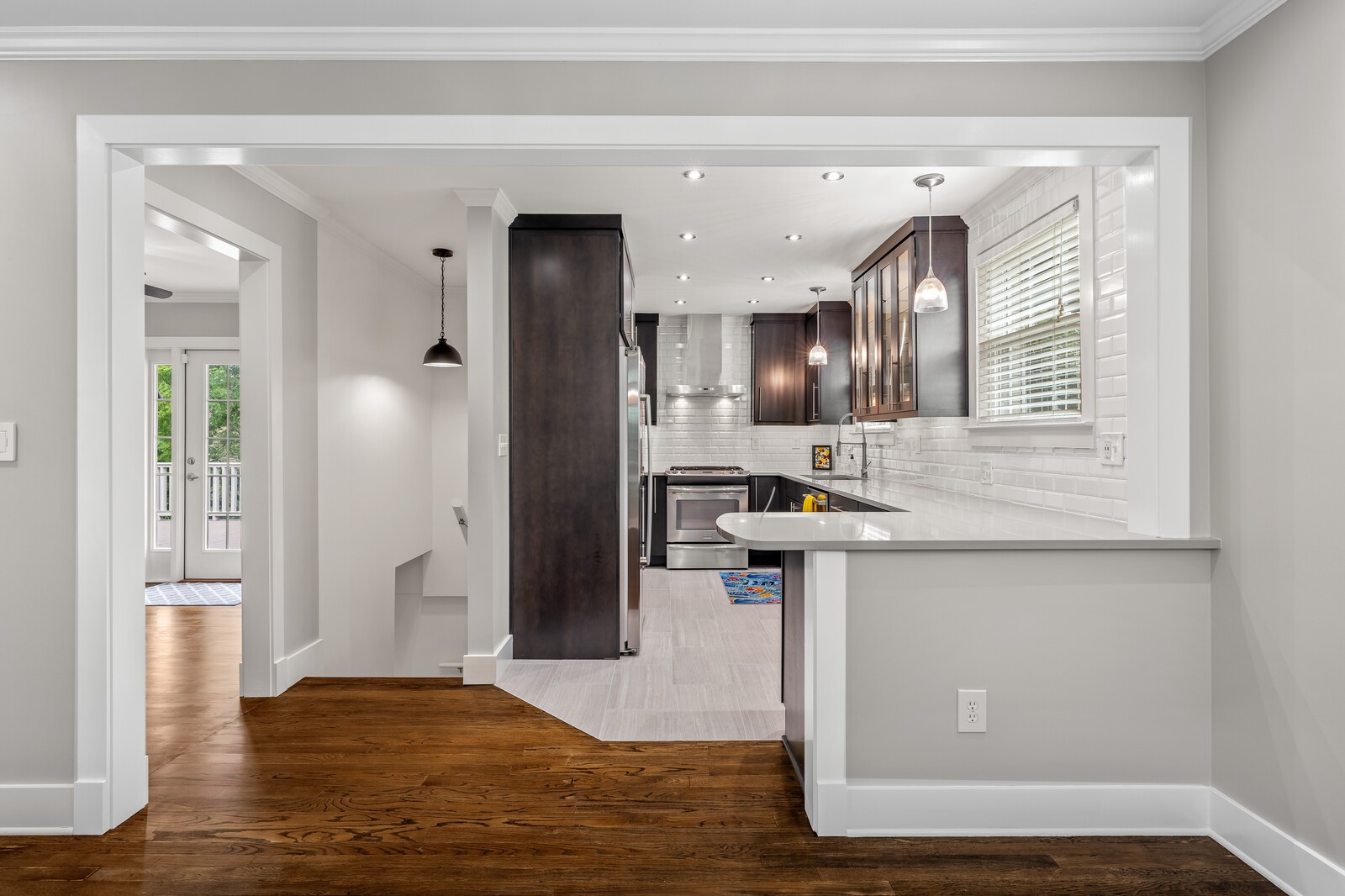 Kitchen with dark cabinets, white tile backsplash, stainless appliances, and a peninsula opening to hardwood floors.