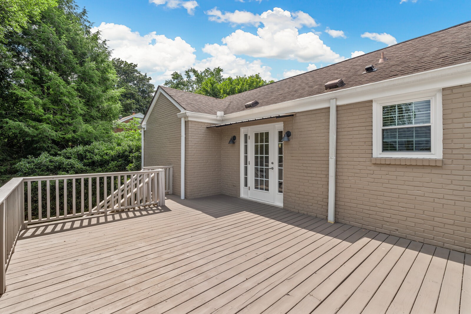 Wide wood deck beside a tan brick house, with white French doors and leafy trees