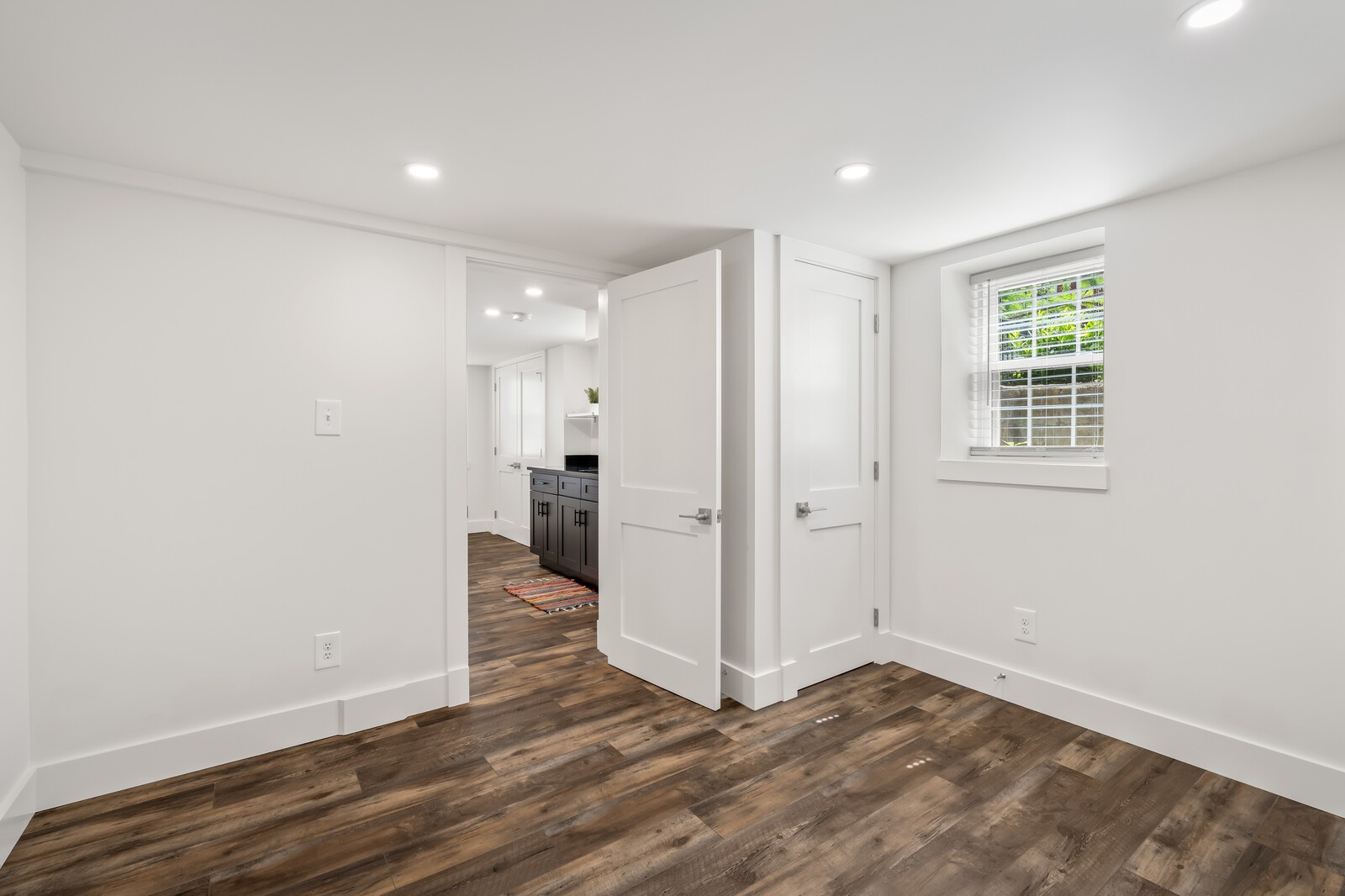 Empty room with dark plank flooring, recessed lights, and a window overlooking greenery.