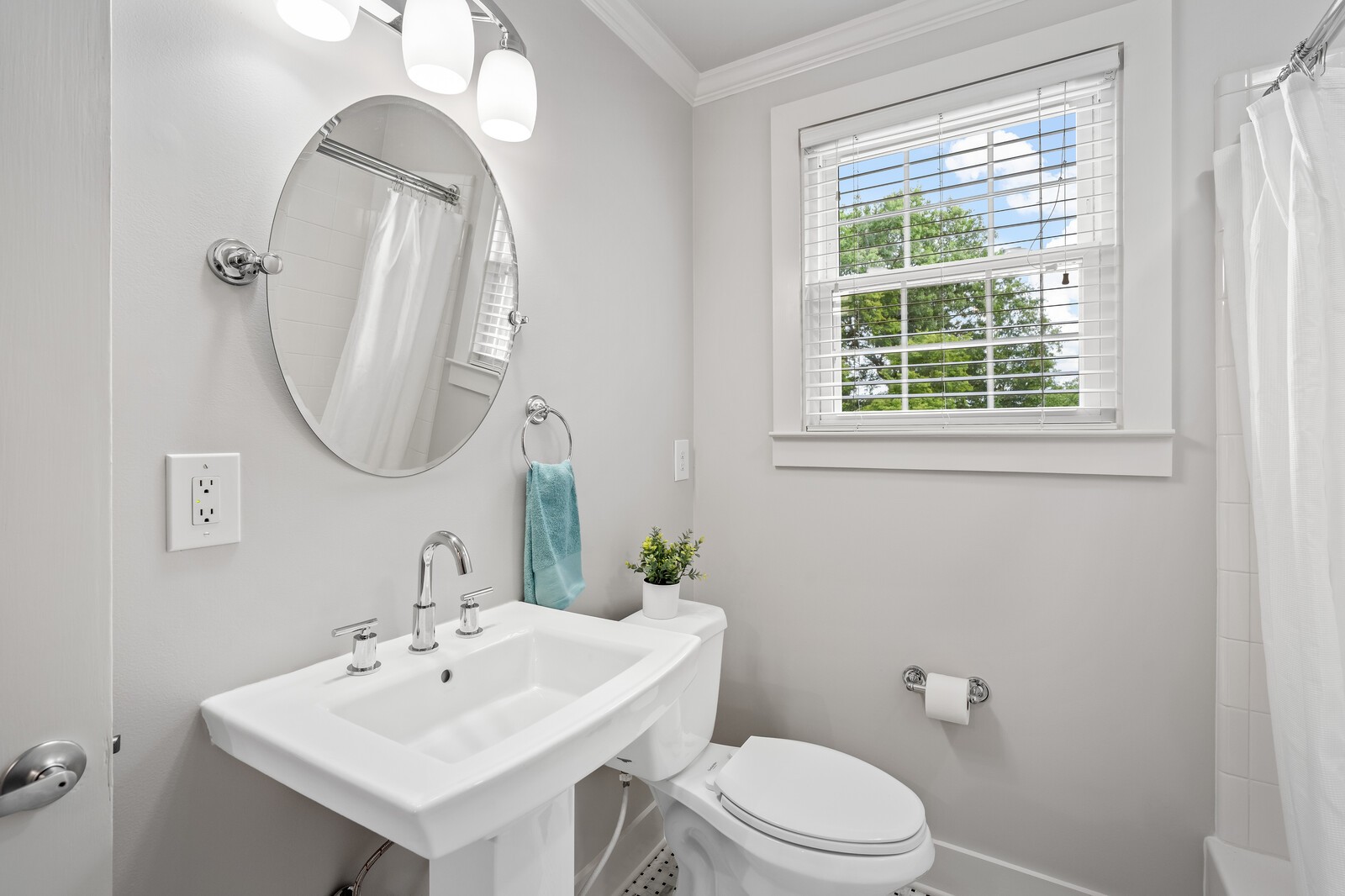 Bathroom with pedestal sink, oval mirror, toilet, and a window overlooking trees.