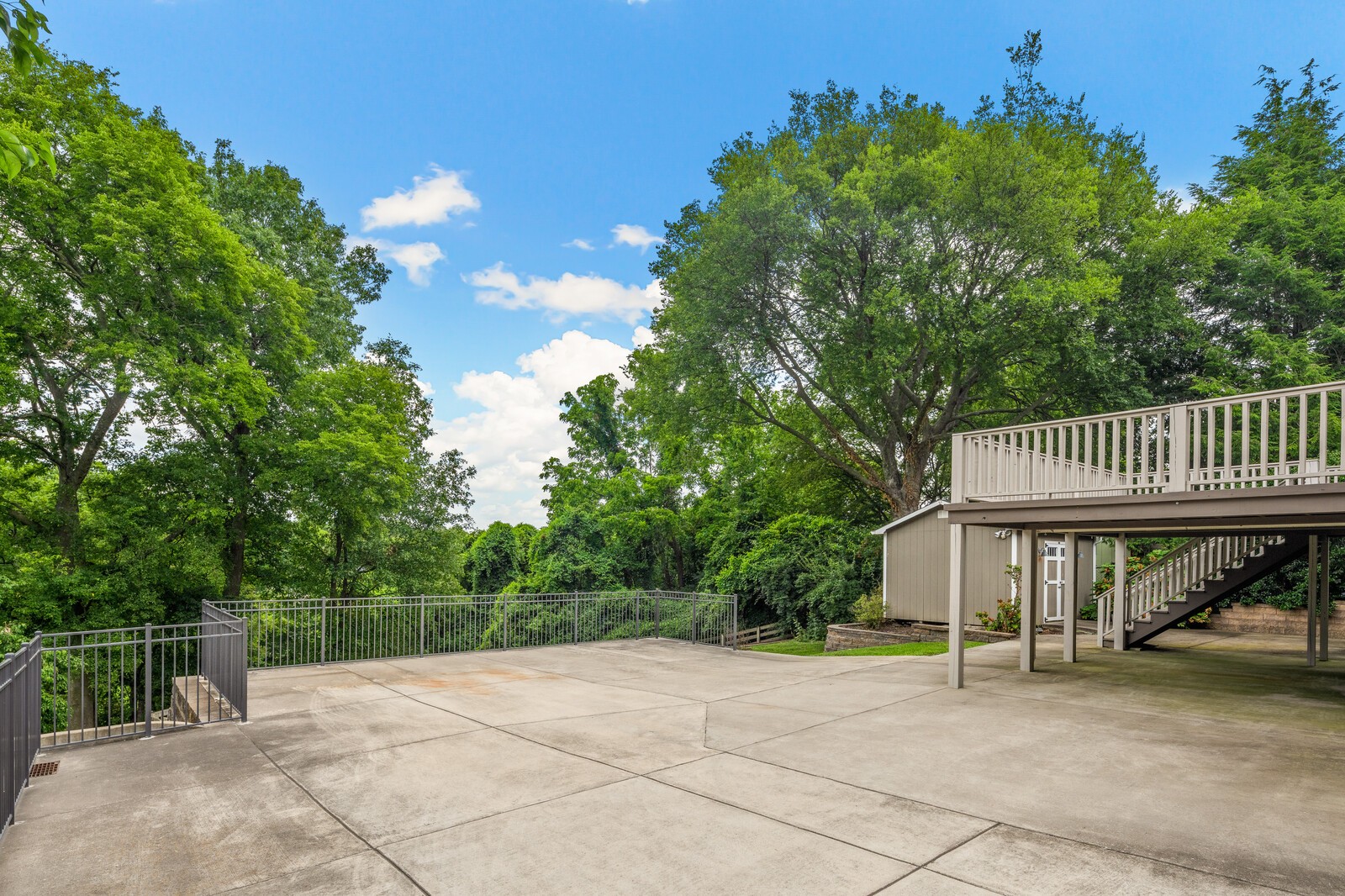Concrete backyard patio beside an elevated deck with white railings, surrounded by mature trees.
