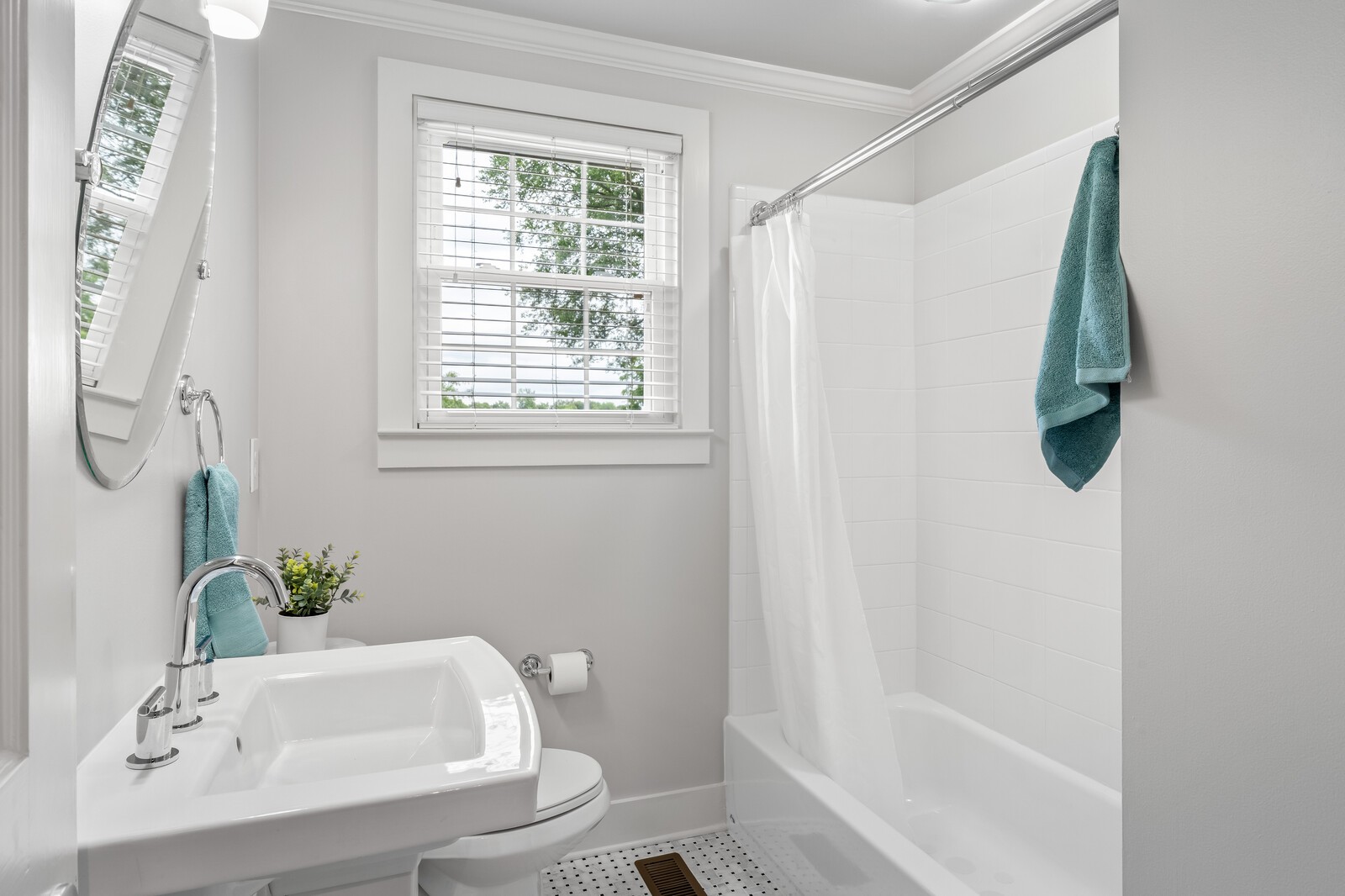 Bright bathroom with a white tub and shower, pedestal sink, and window with blinds.