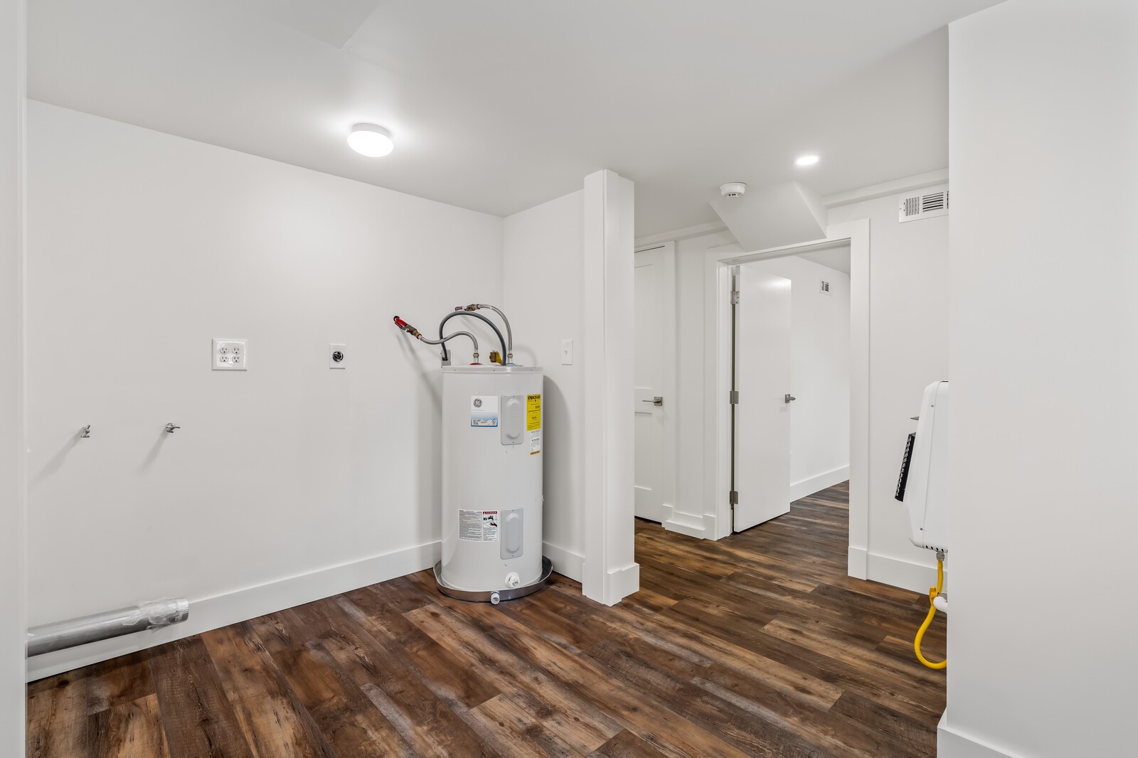 Utility room with a water heater, exposed plumbing connections, and dark wood-look flooring.