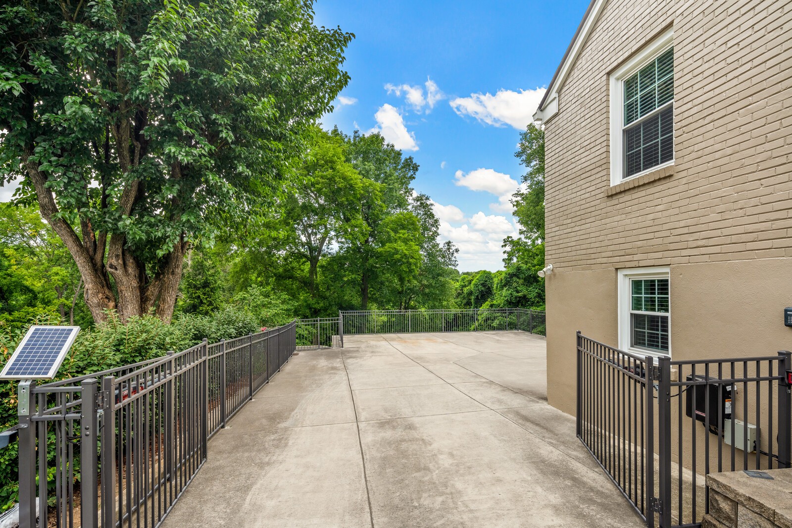 Concrete patio beside a brick home, enclosed by black railings and shaded by mature trees.