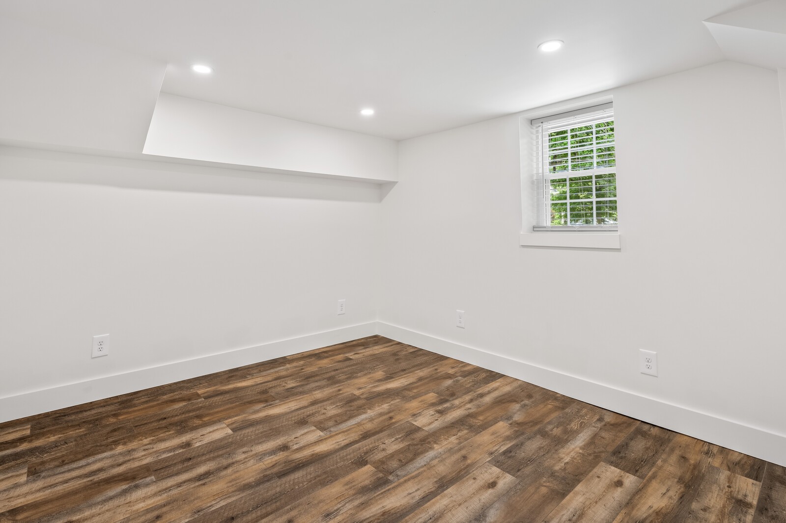 Empty room with brown plank flooring, white walls, recessed lights, and a small window with blinds.