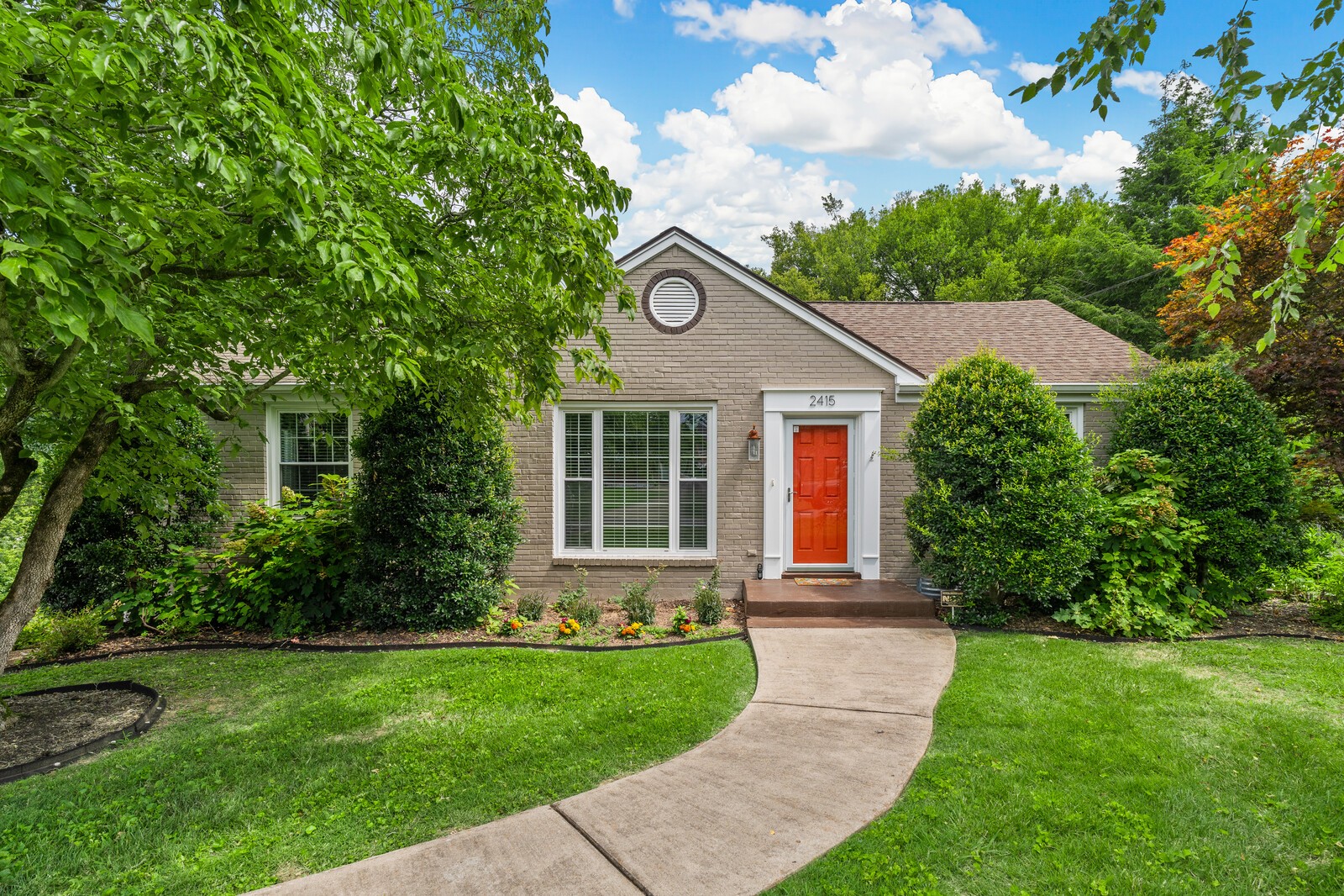 Taupe brick home with a red front door, mature landscaping, and a curved walkway.