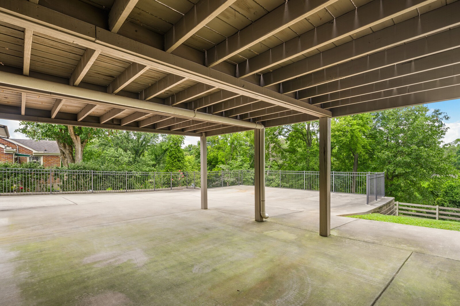 Covered concrete patio beneath a raised wood deck, with metal railings and leafy trees beyond.