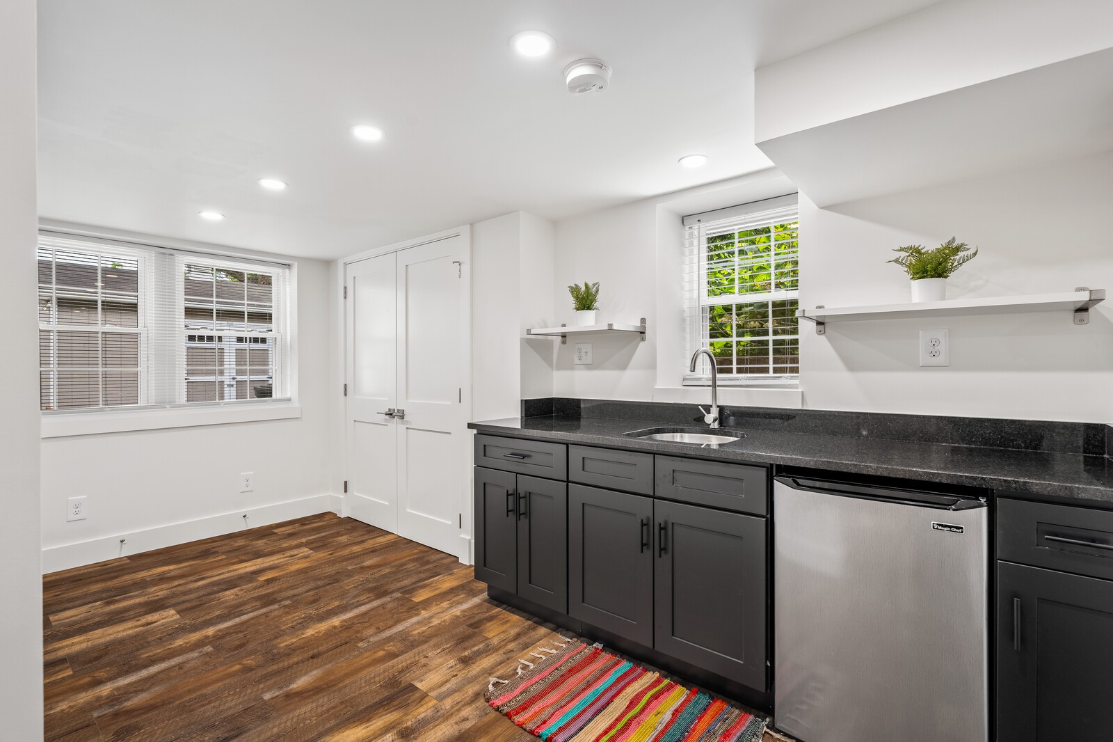 Kitchen with dark cabinets, black countertops, stainless steel dishwasher, wood flooring, and a colorful woven rug.