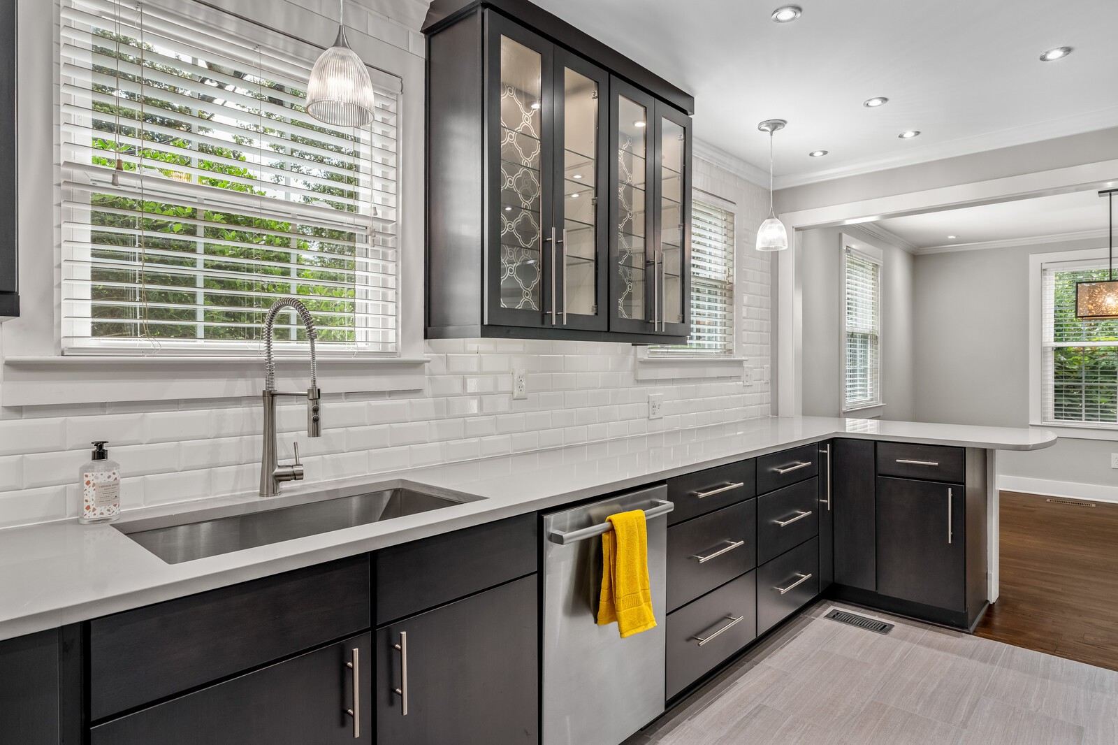 Kitchen with dark cabinets, white counters and tile, stainless sink, and large windows.