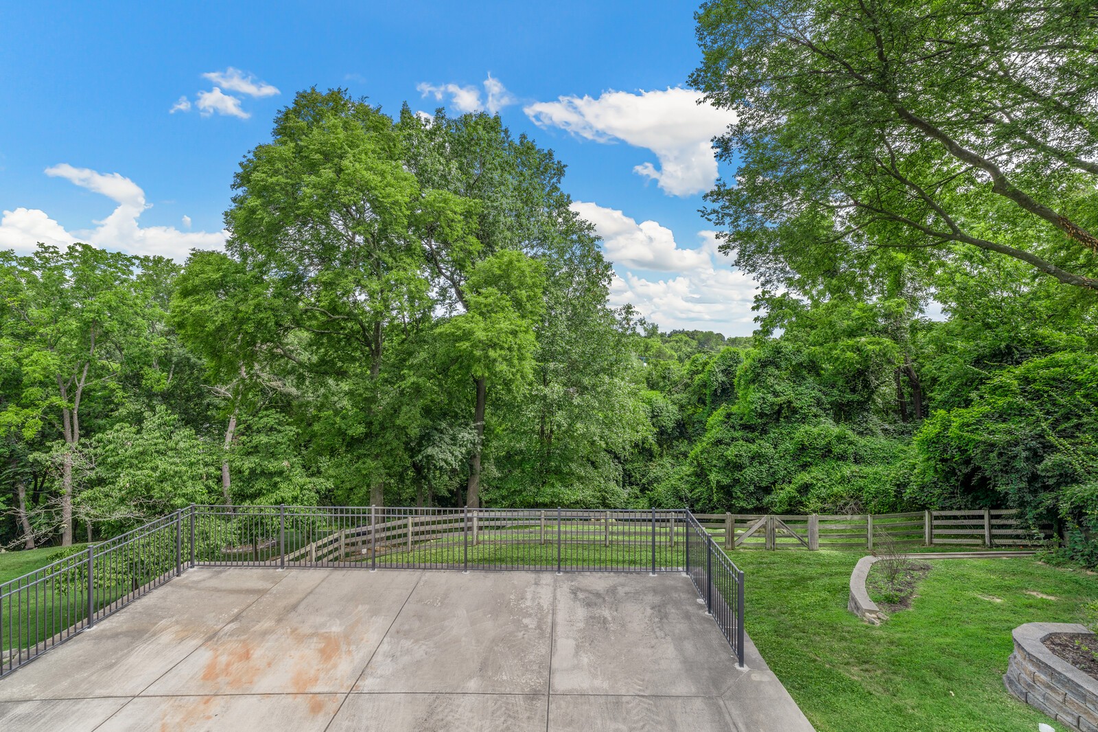 Concrete patio with black railing overlooking a fenced, tree-lined backyard.