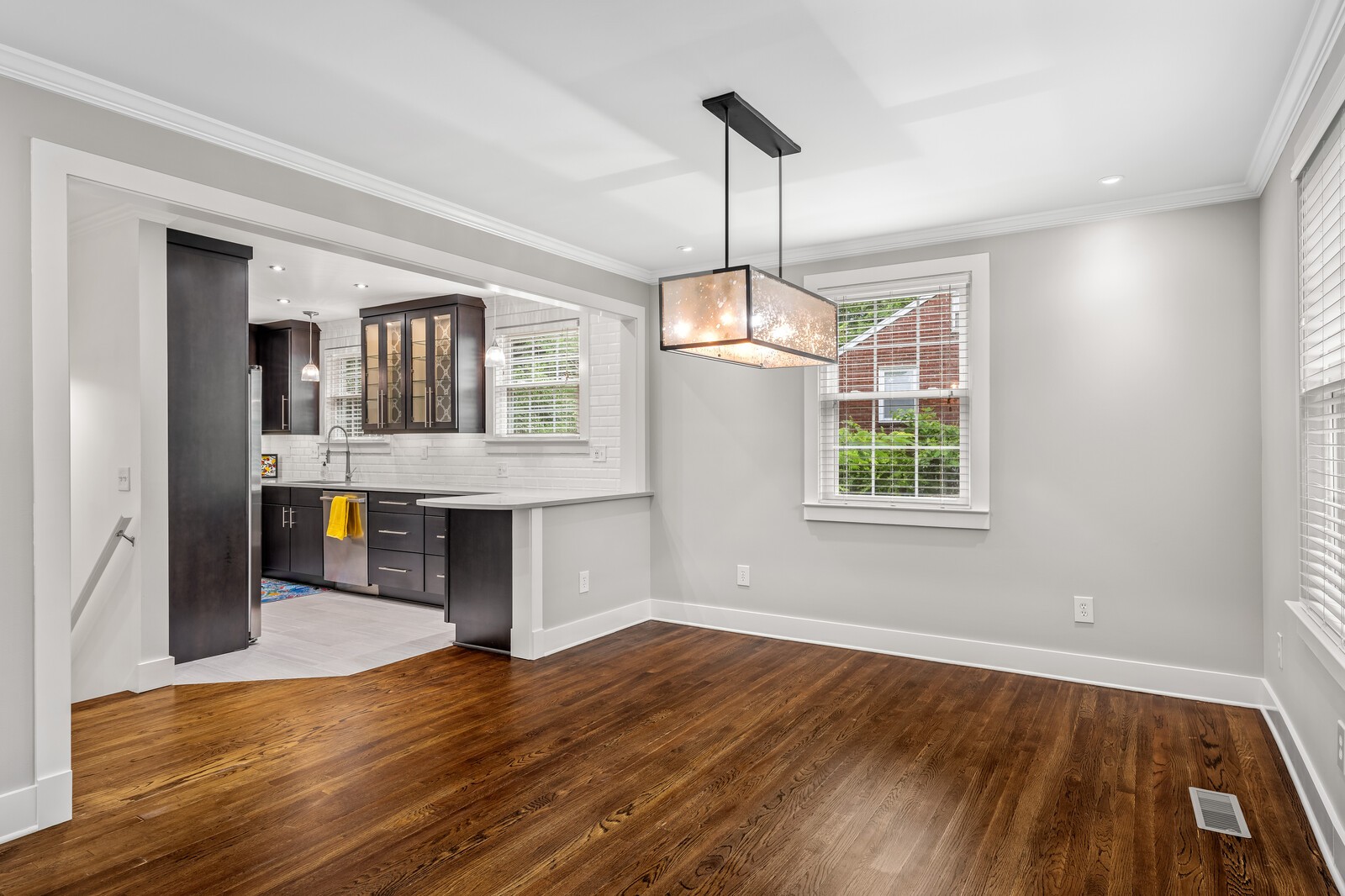 Empty dining room with hardwood floors and pendant light, open to a dark-cabinet kitchen.