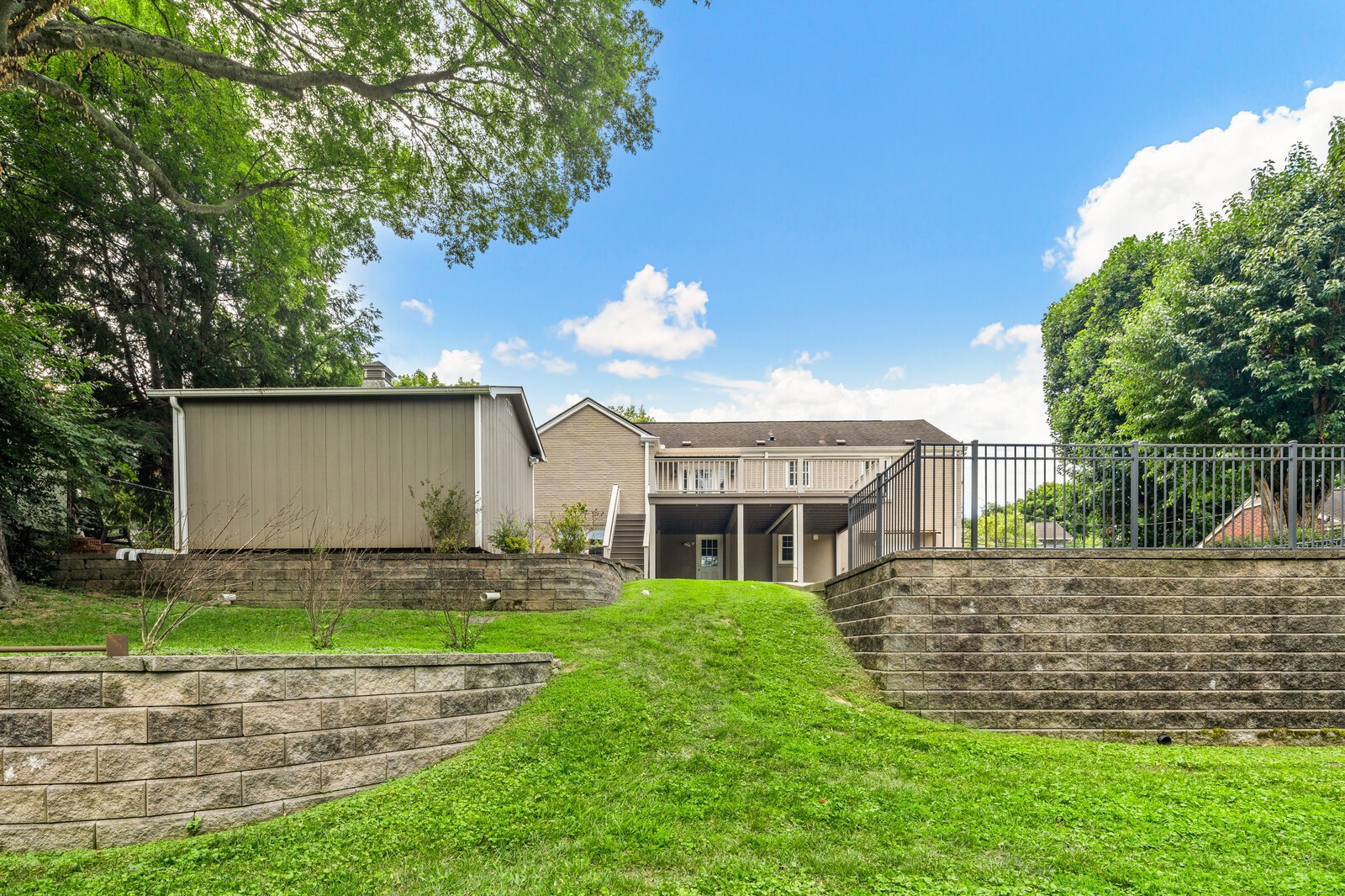 Home’s rear exterior with an elevated deck, grassy sloped yard, and stone retaining walls.