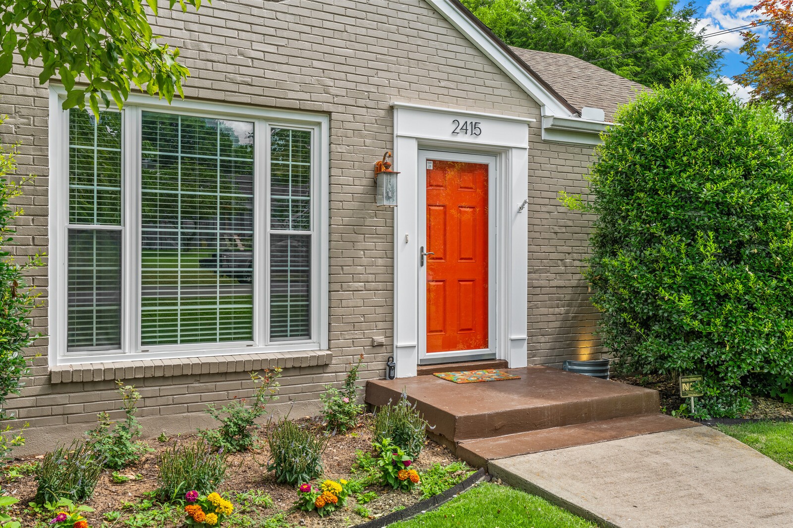 Brick home exterior with a bright orange-red front door, large window, and landscaped entry.