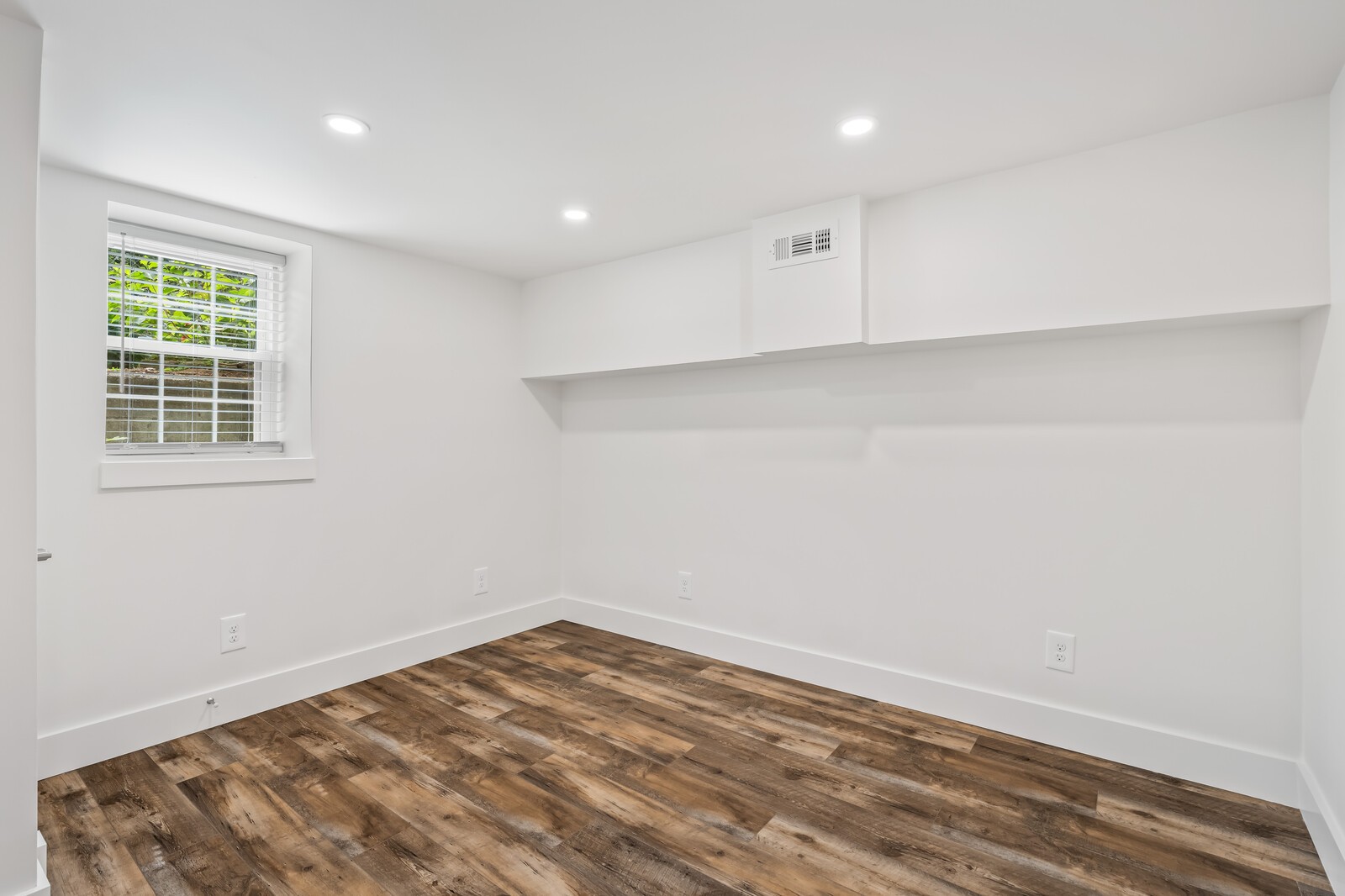 Empty room with brown wood-look flooring, a window with blinds, and a wide wall soffit.