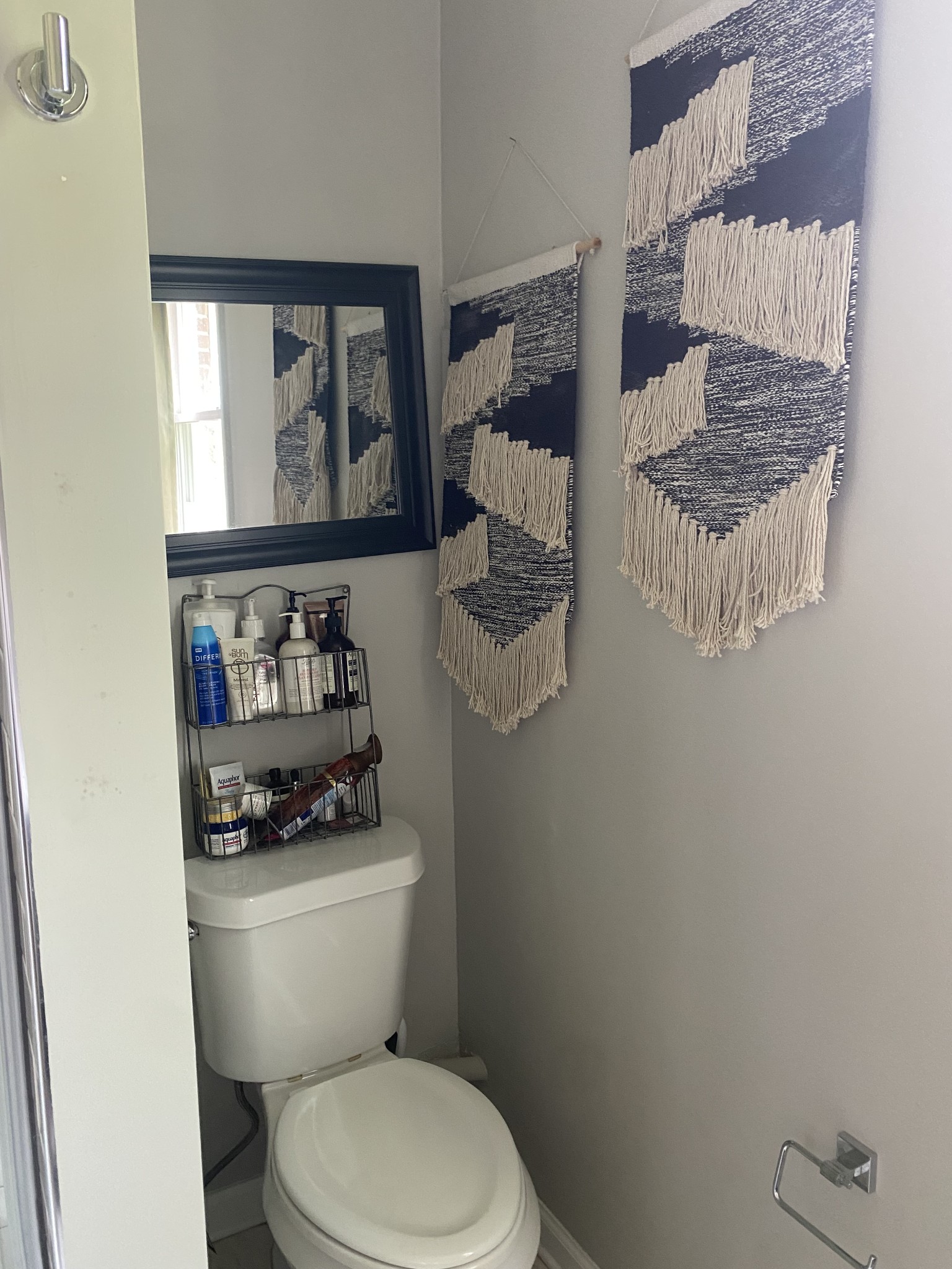 Bathroom toilet nook with a black-framed mirror, toiletries, and woven wall hangings.