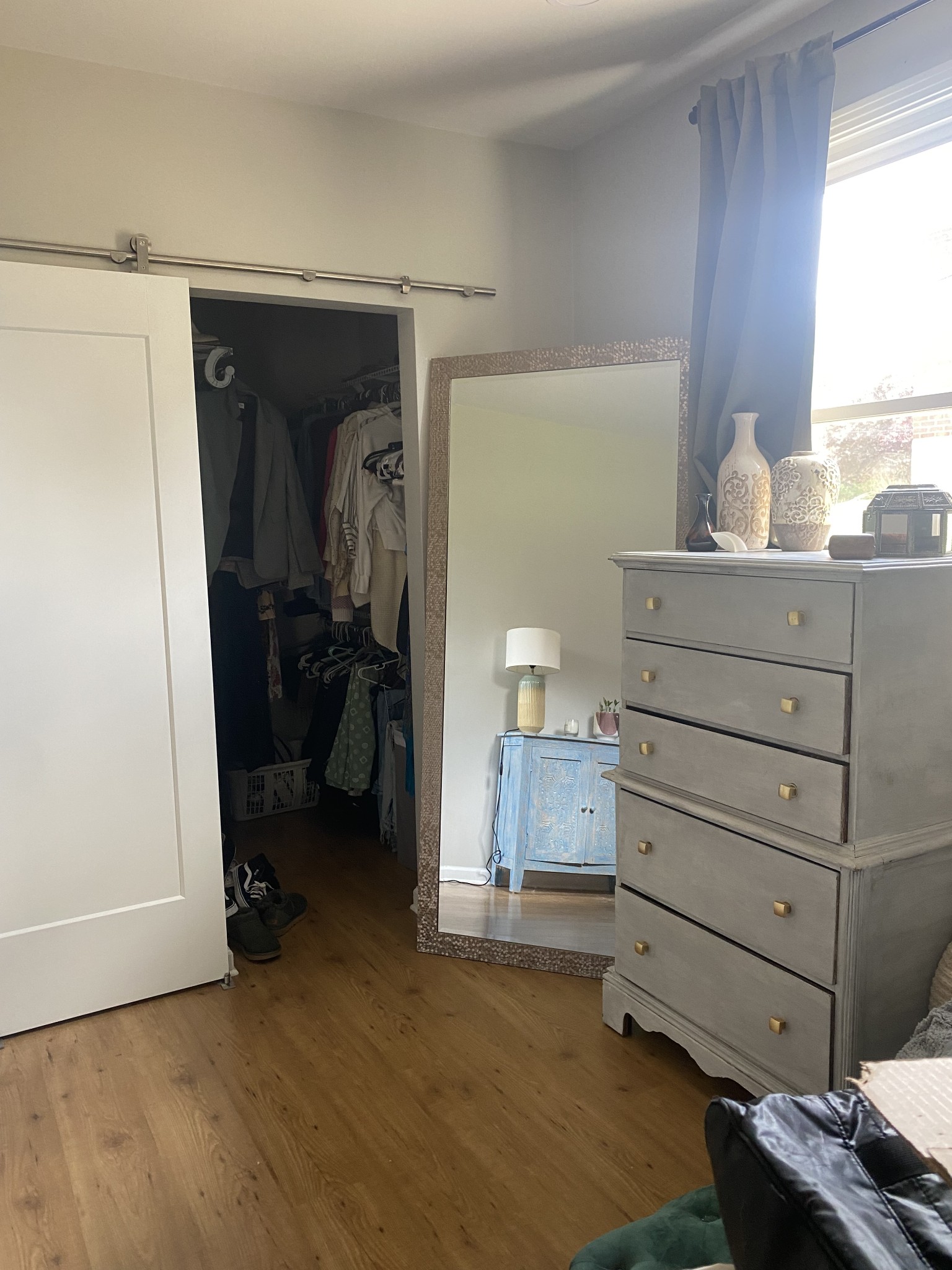 Bedroom with an open clothing-filled closet, leaning mirror, gray dresser, and hardwood floors.