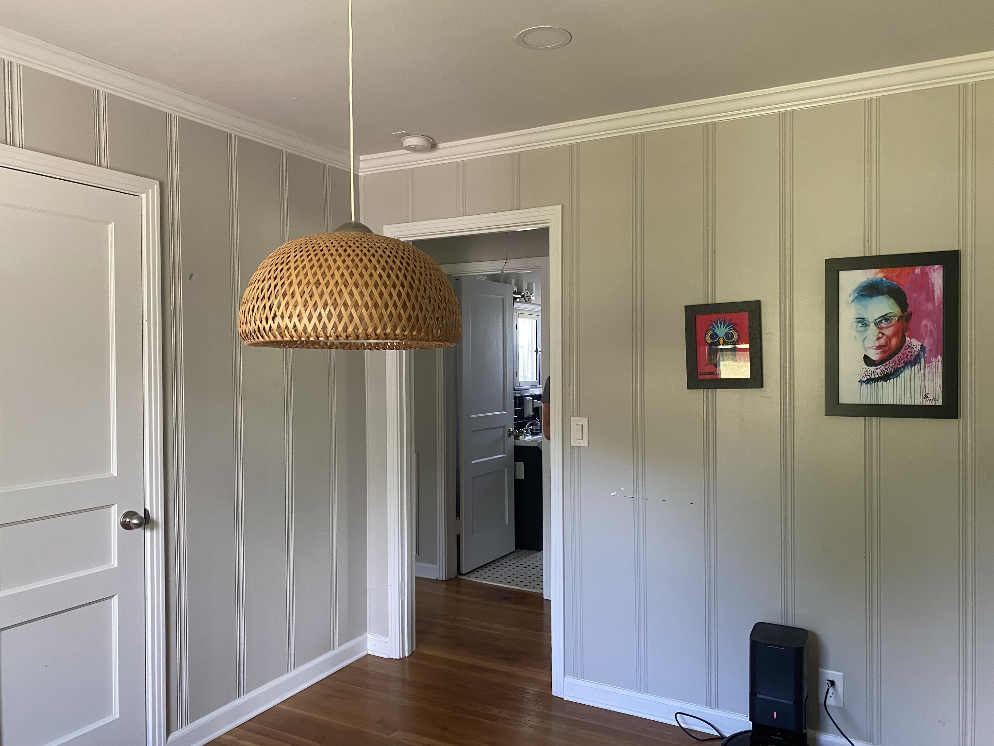 Paneled room with hardwood floors, a woven pendant light, framed artwork, and an open doorway.