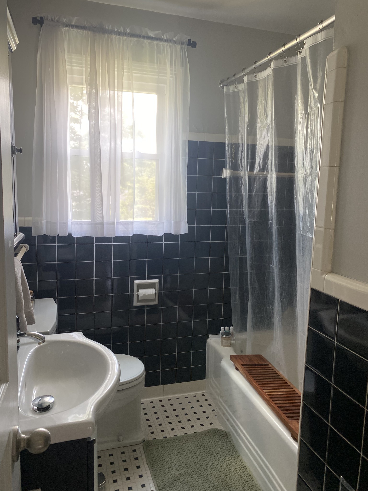 Bathroom with navy tile, white bathtub, wooden bath tray, and a bright window with sheer curtains.
