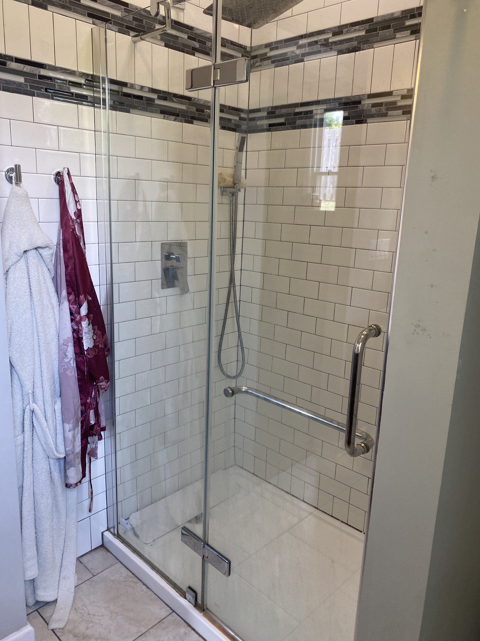 Walk-in shower with glass doors, white subway tile, dark mosaic accent band, and chrome grab bar.