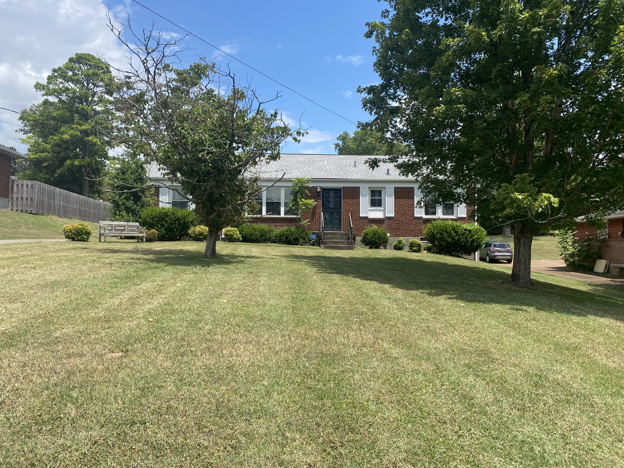Brick ranch home with a broad sloping lawn, mature trees, and a wooden bench.