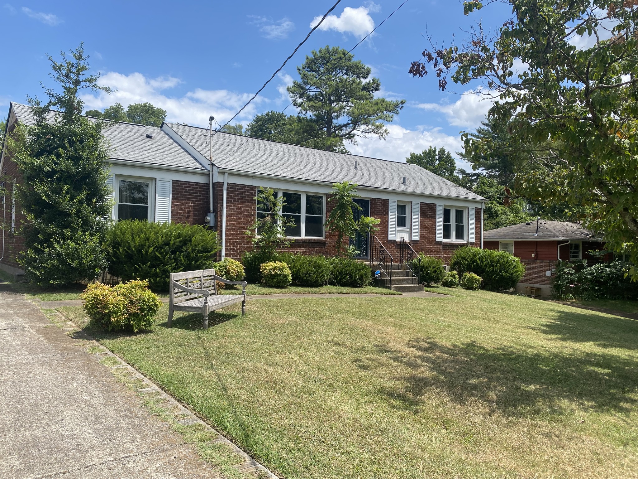 Red brick ranch house with white shutters, front steps, mature shrubs, and a wooden bench on the lawn.