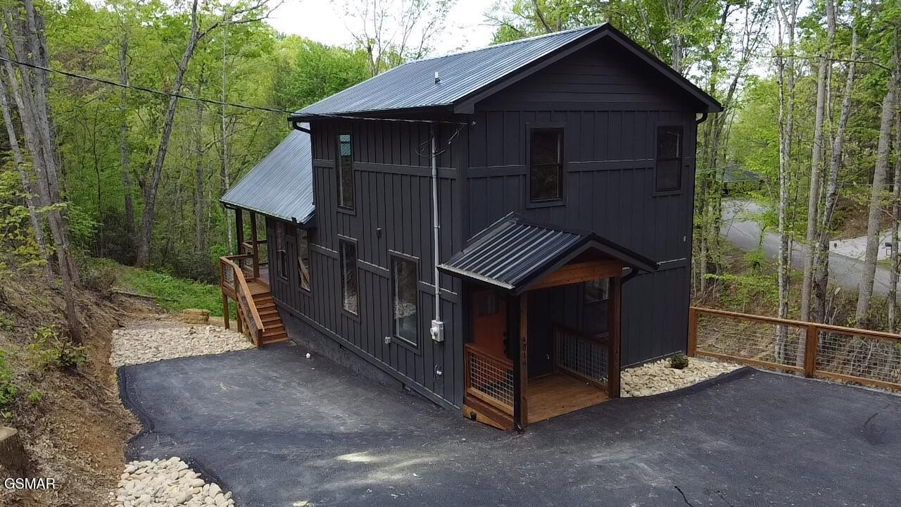 Dark two-story home with metal roofs, a covered wooden porch, and a paved driveway among trees.