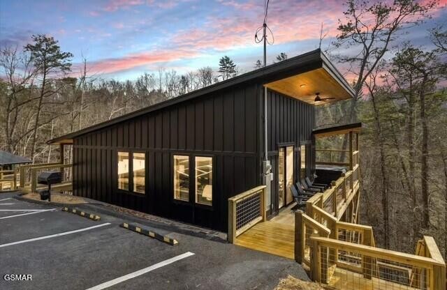 Modern dark-sided home with a raised wooden deck, parking area, and wooded surroundings at dusk.