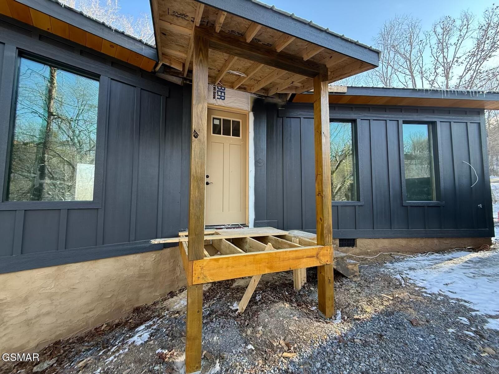 Dark gray home with an unfinished timber porch, light front door, and snow on the ground.
