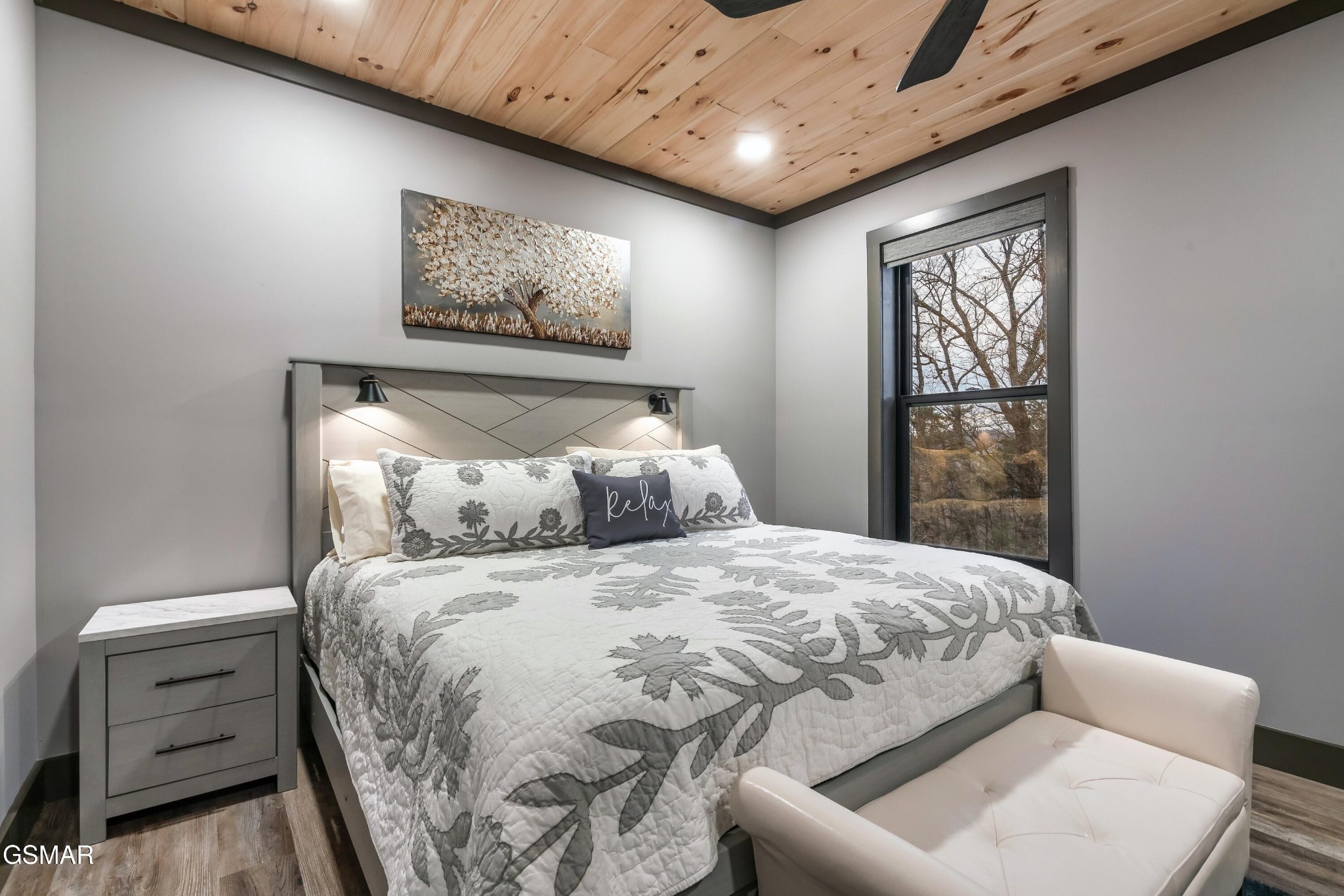Bedroom with a floral quilted bed, wood ceiling, and tall window overlooking bare trees.