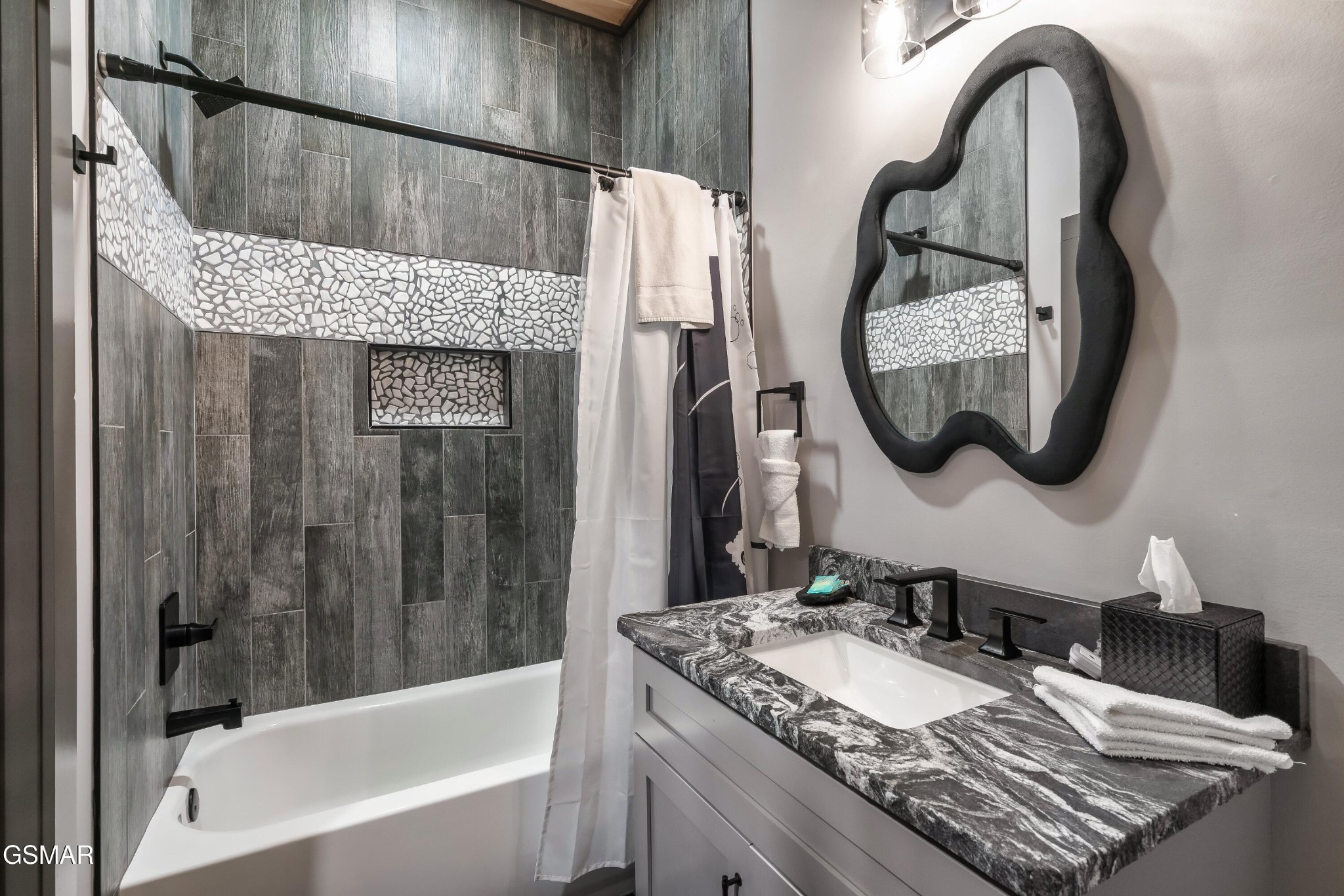 Bathroom with dark tile shower, mosaic accent band, stone vanity countertop, and wavy black-framed mirror.