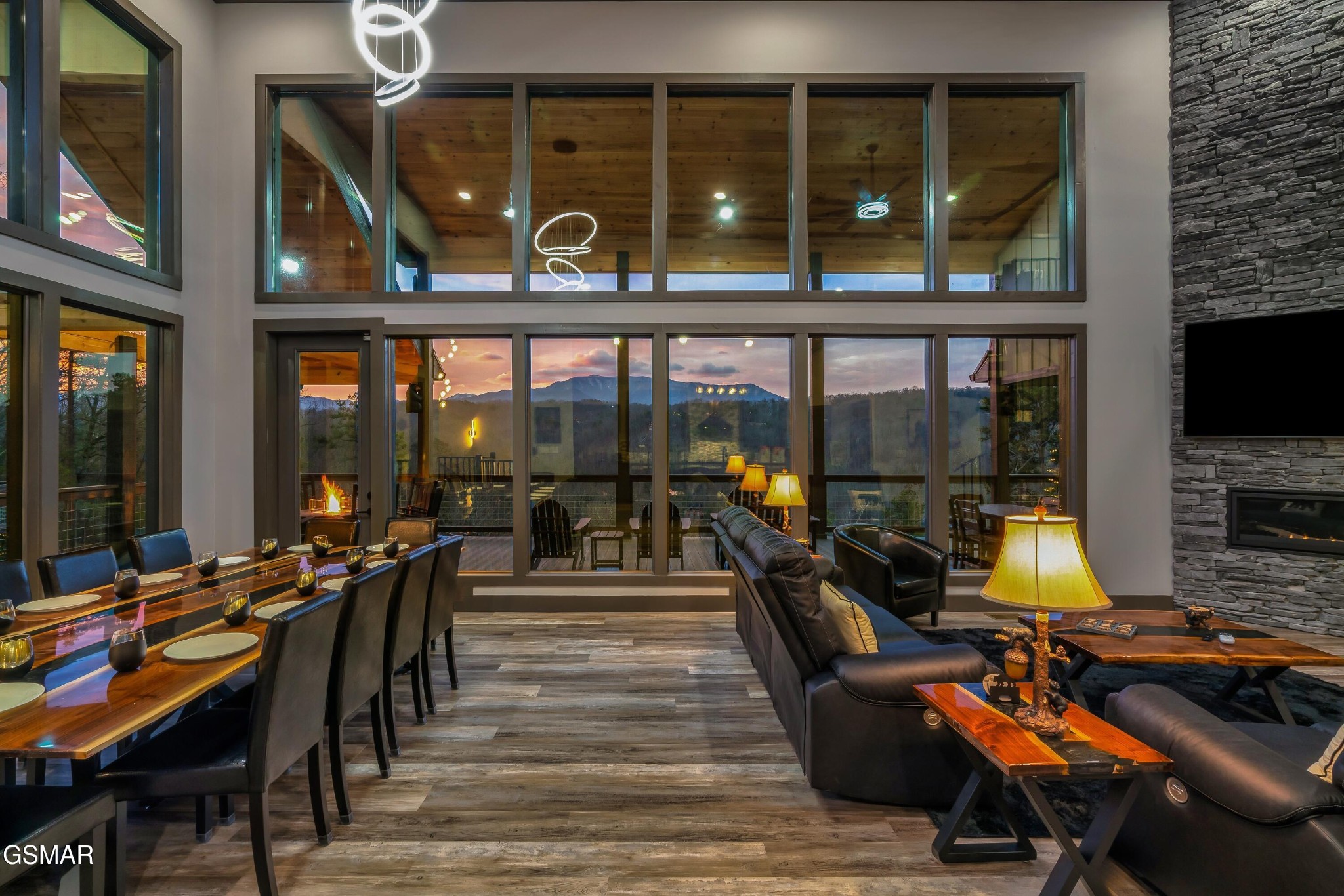 Open-plan living and dining room with leather seating, a stone fireplace, and sunset mountain views through tall windows.