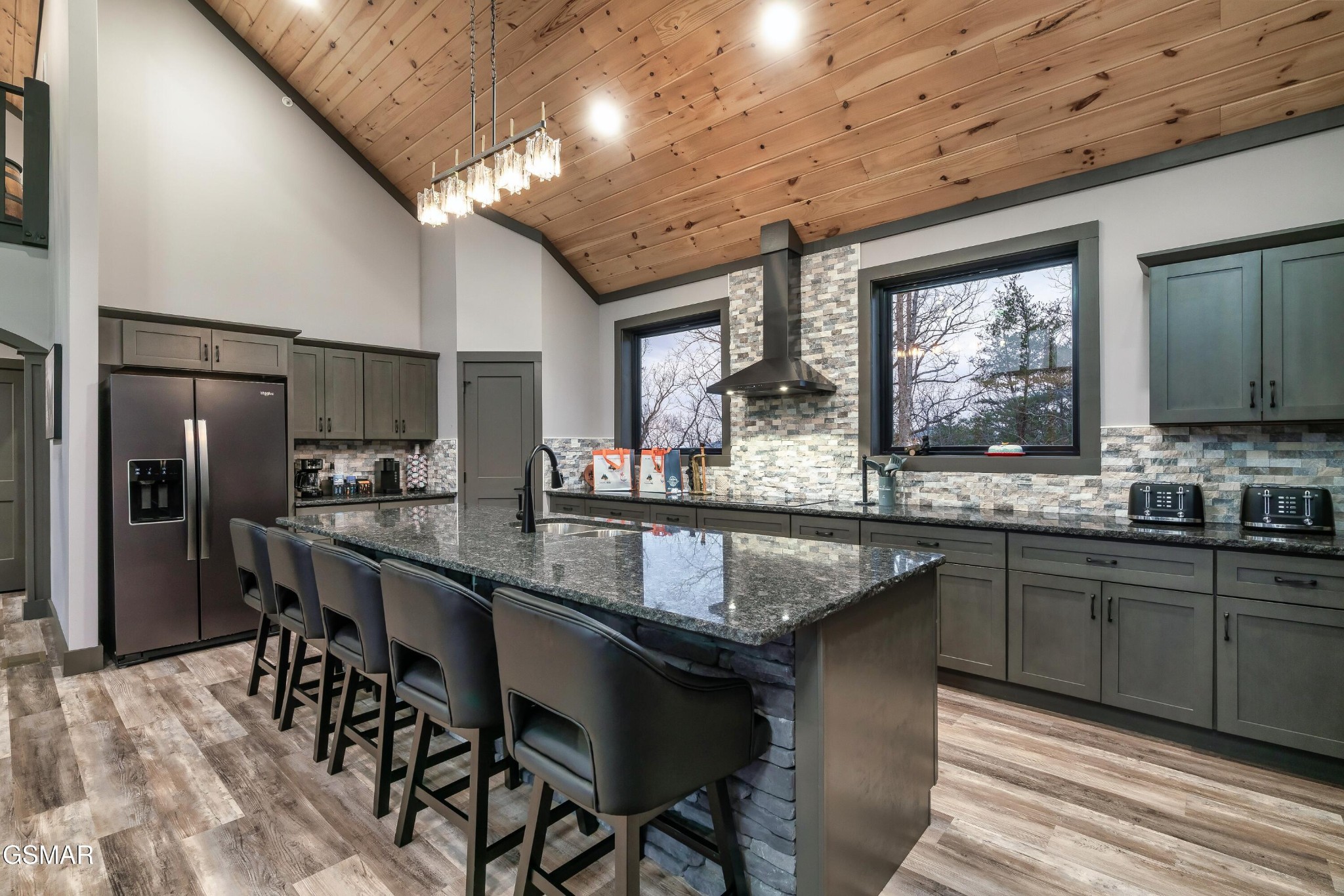 Modern kitchen with a large island, dark cabinets, stone backsplash, and vaulted wood ceiling.