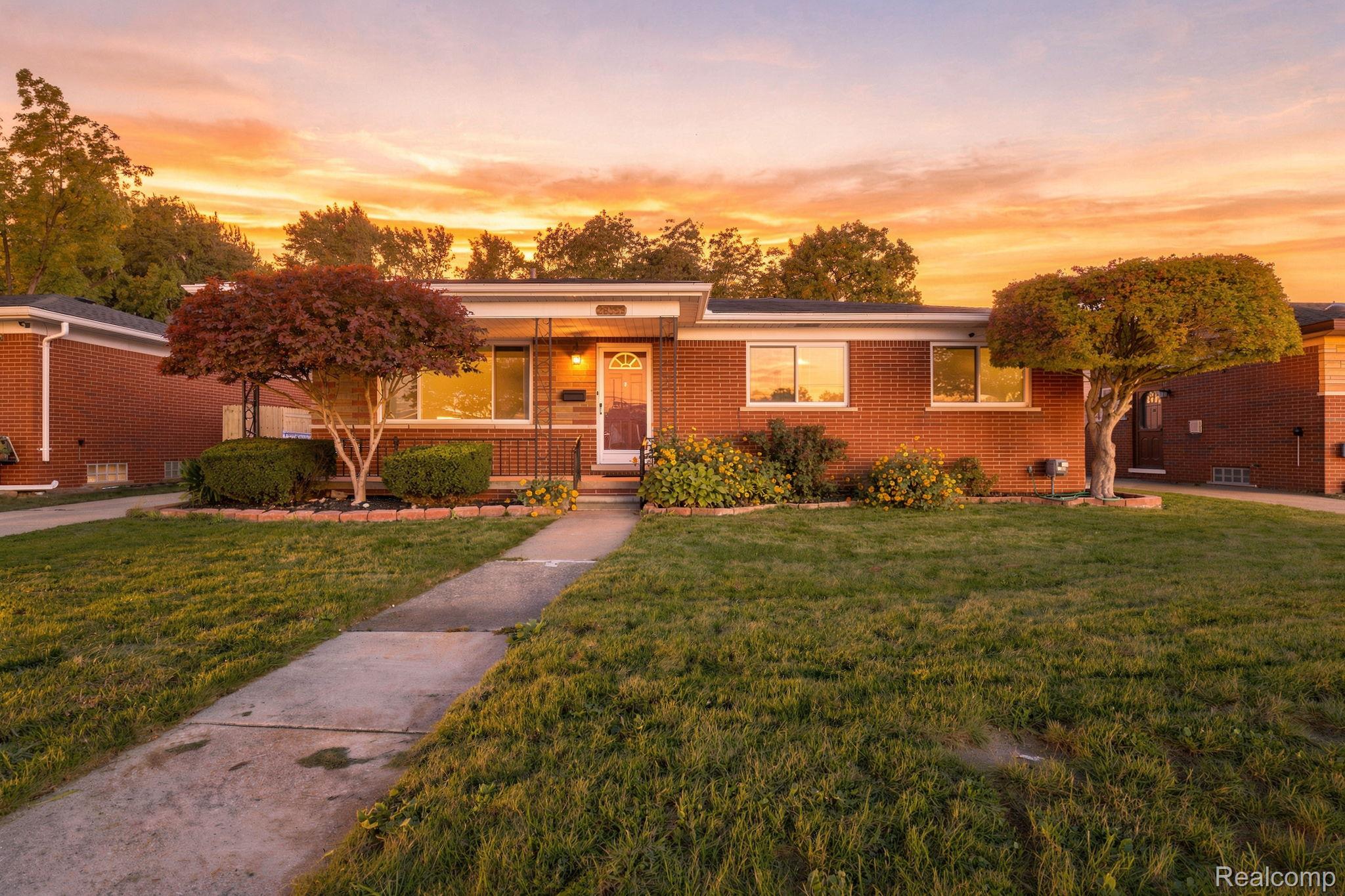 Single-story brick house with flower beds and trimmed trees, photographed at sunset.
