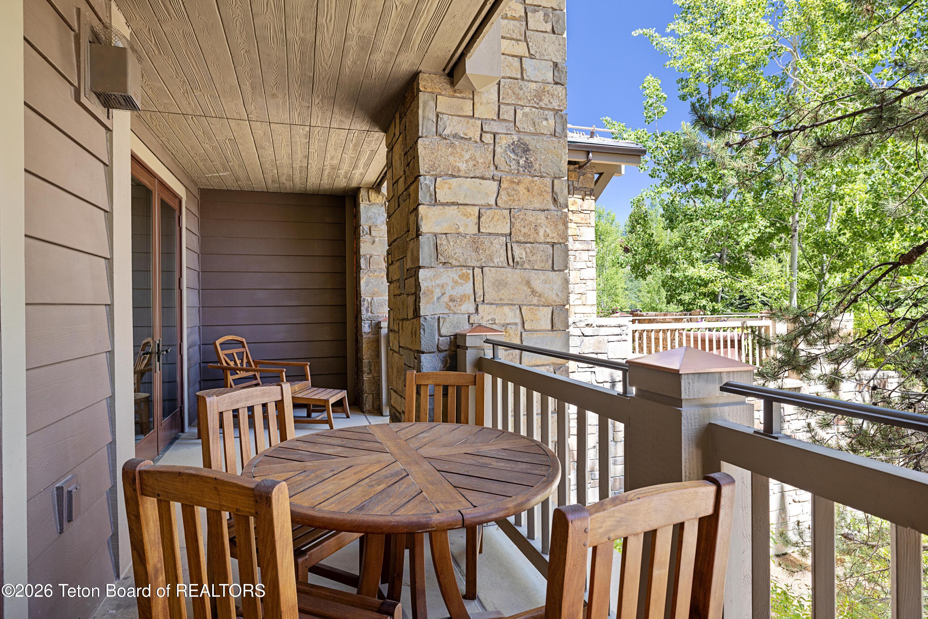 Covered balcony with a round wooden table and chairs beside stone columns and leafy trees.
