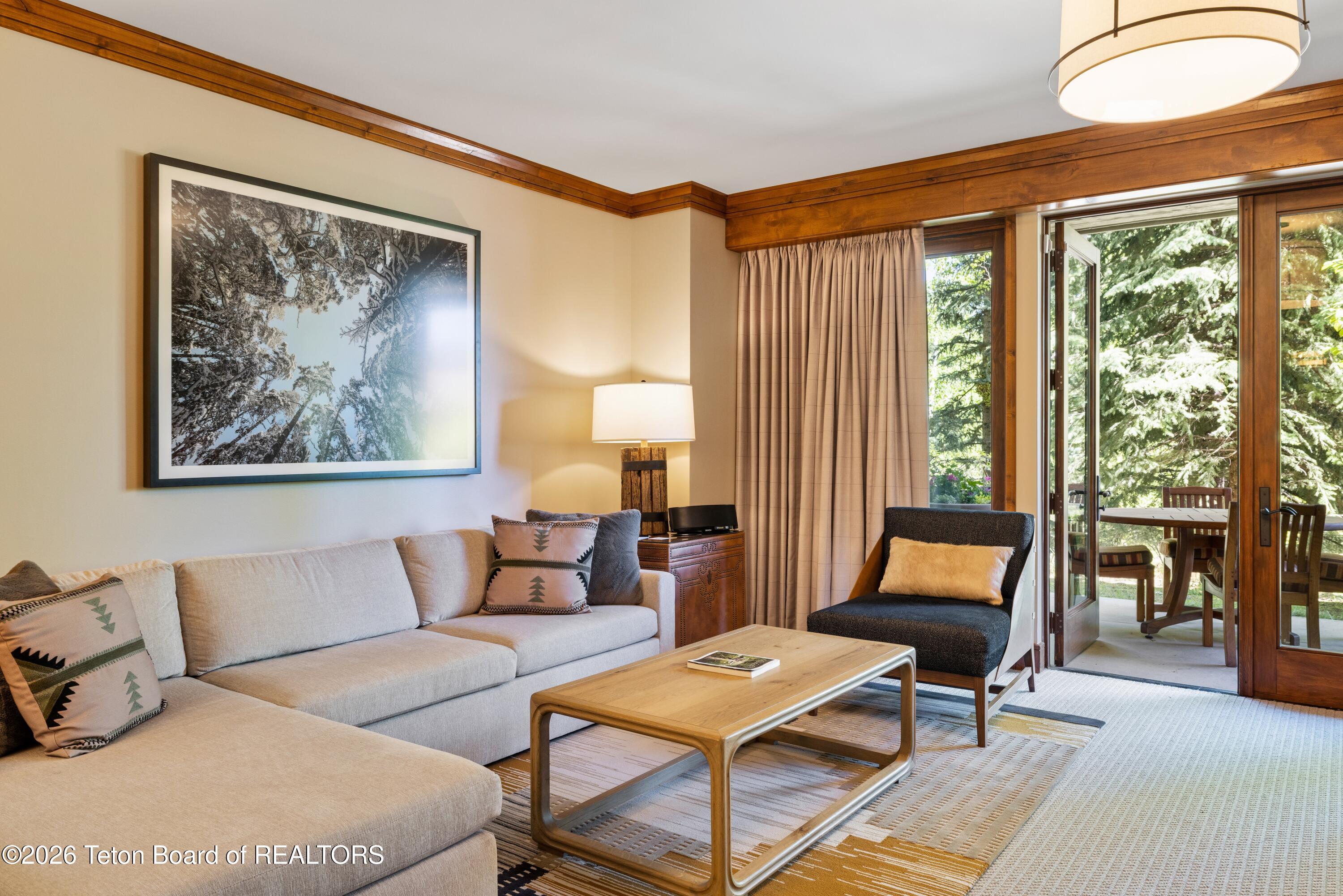 Living room with a light sectional and wood coffee table beside glass doors opening to a tree-lined patio.