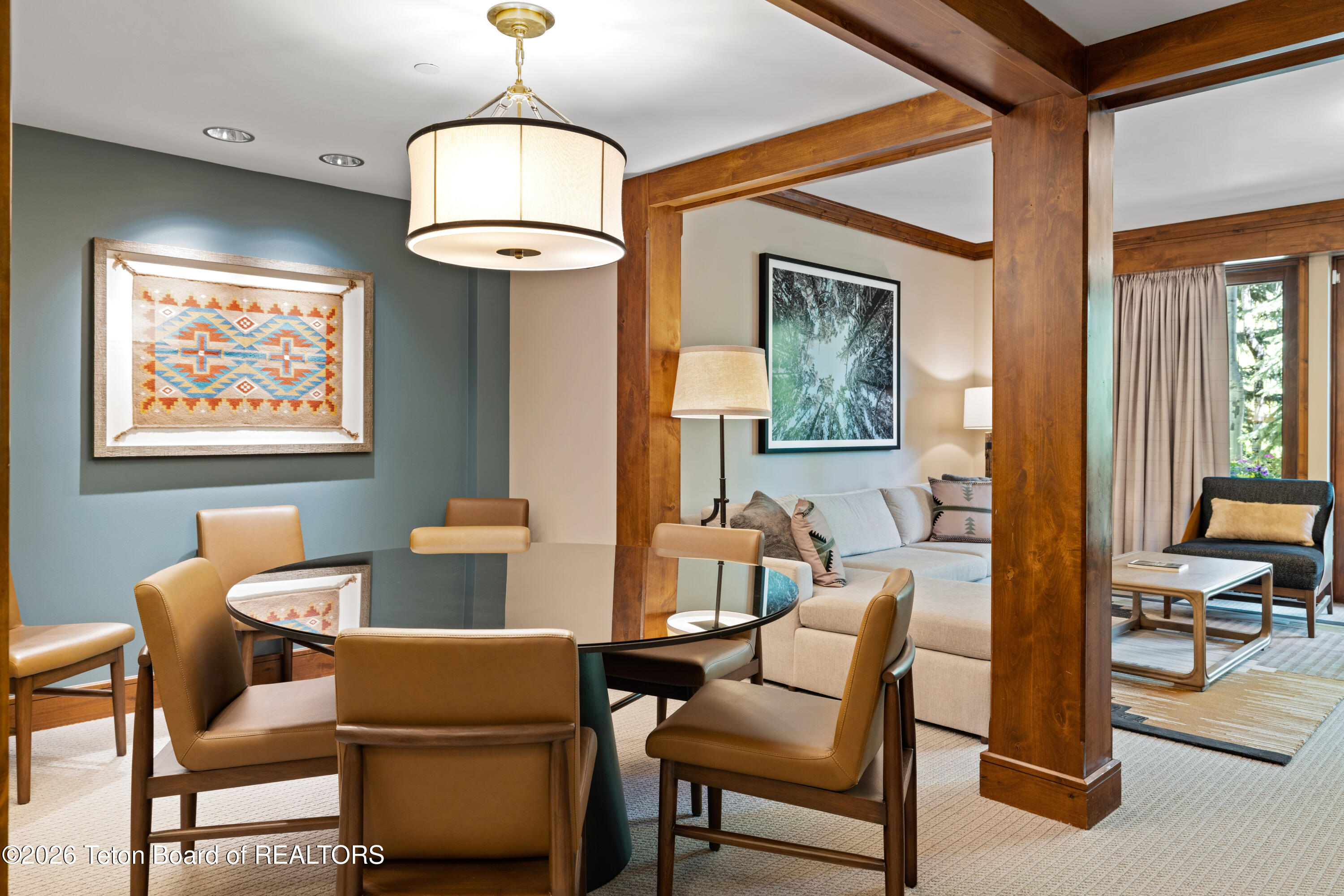 Dining area with a round table and tan leather chairs opens to a living room framed by wood beams.